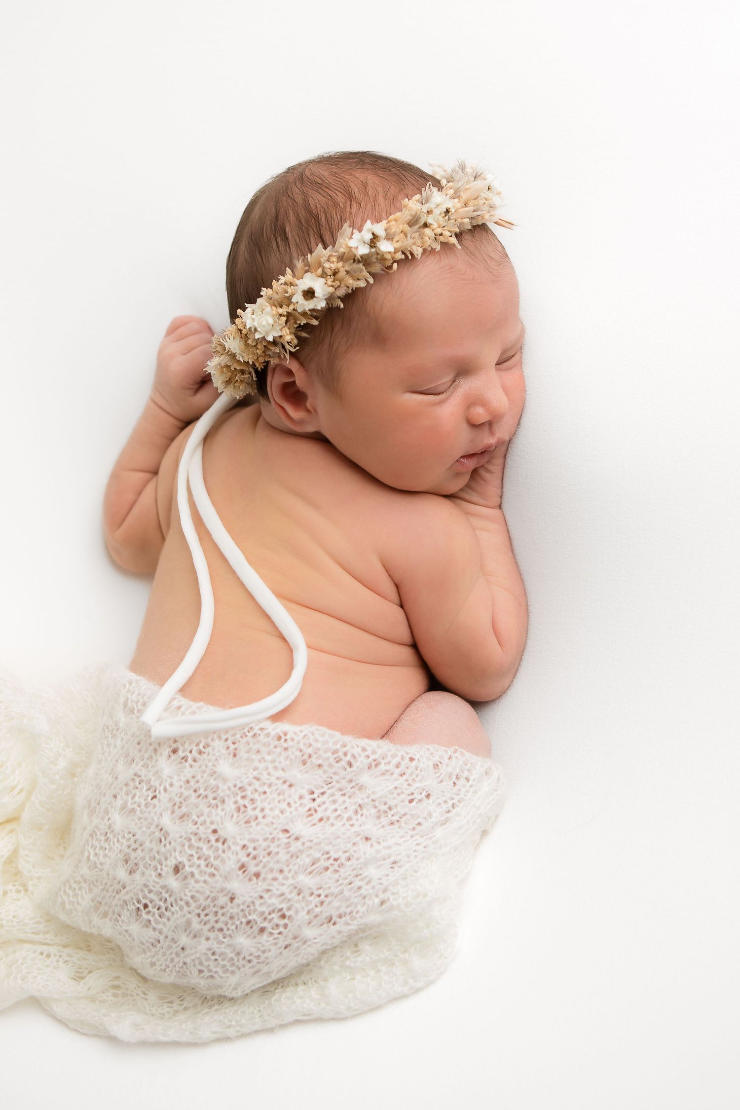 Newborn in lace wrap and floral crown sleeping peacefully on white backdrop.