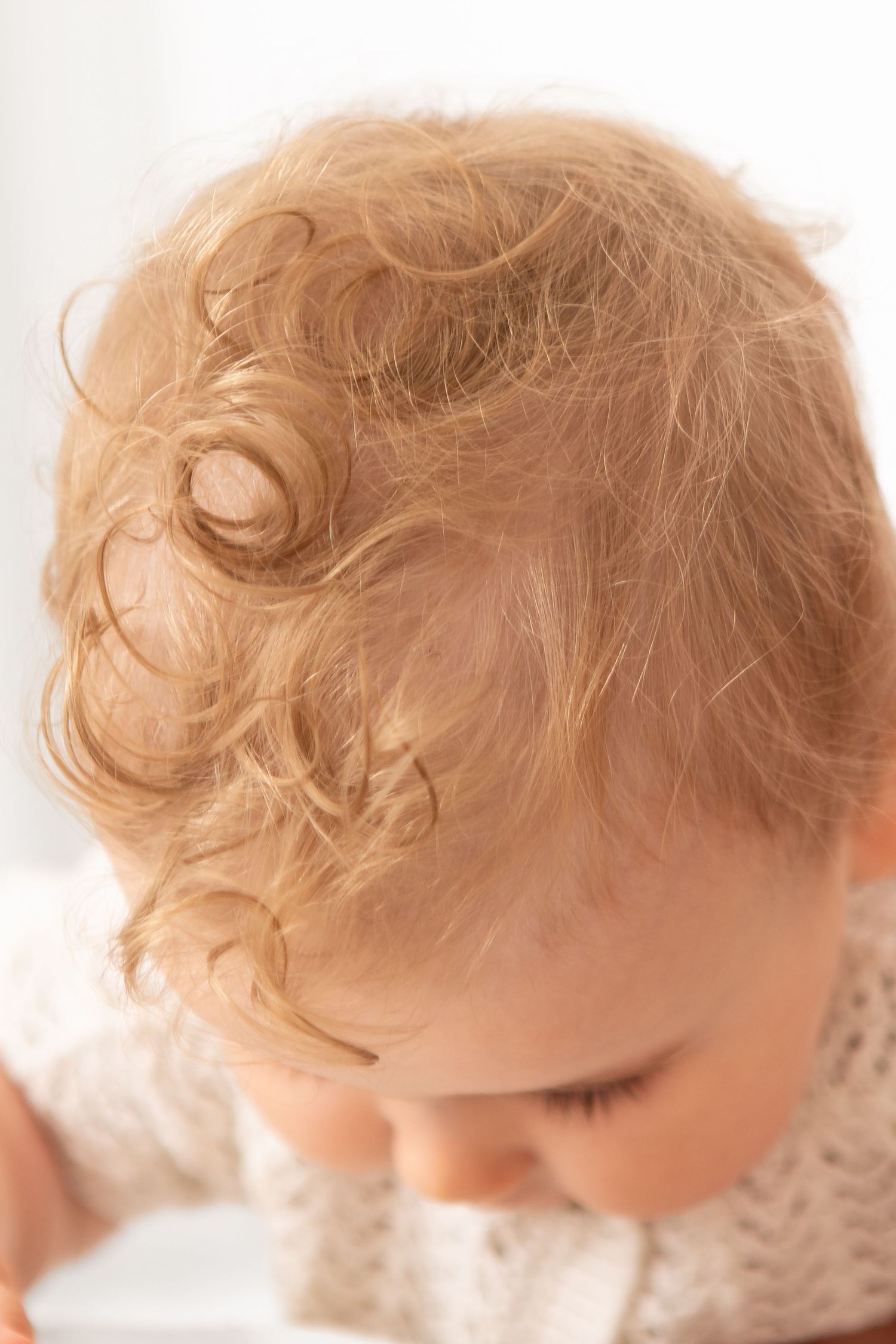 Close-up of a baby with soft curly blonde hair looking down, captured in warm natural light.