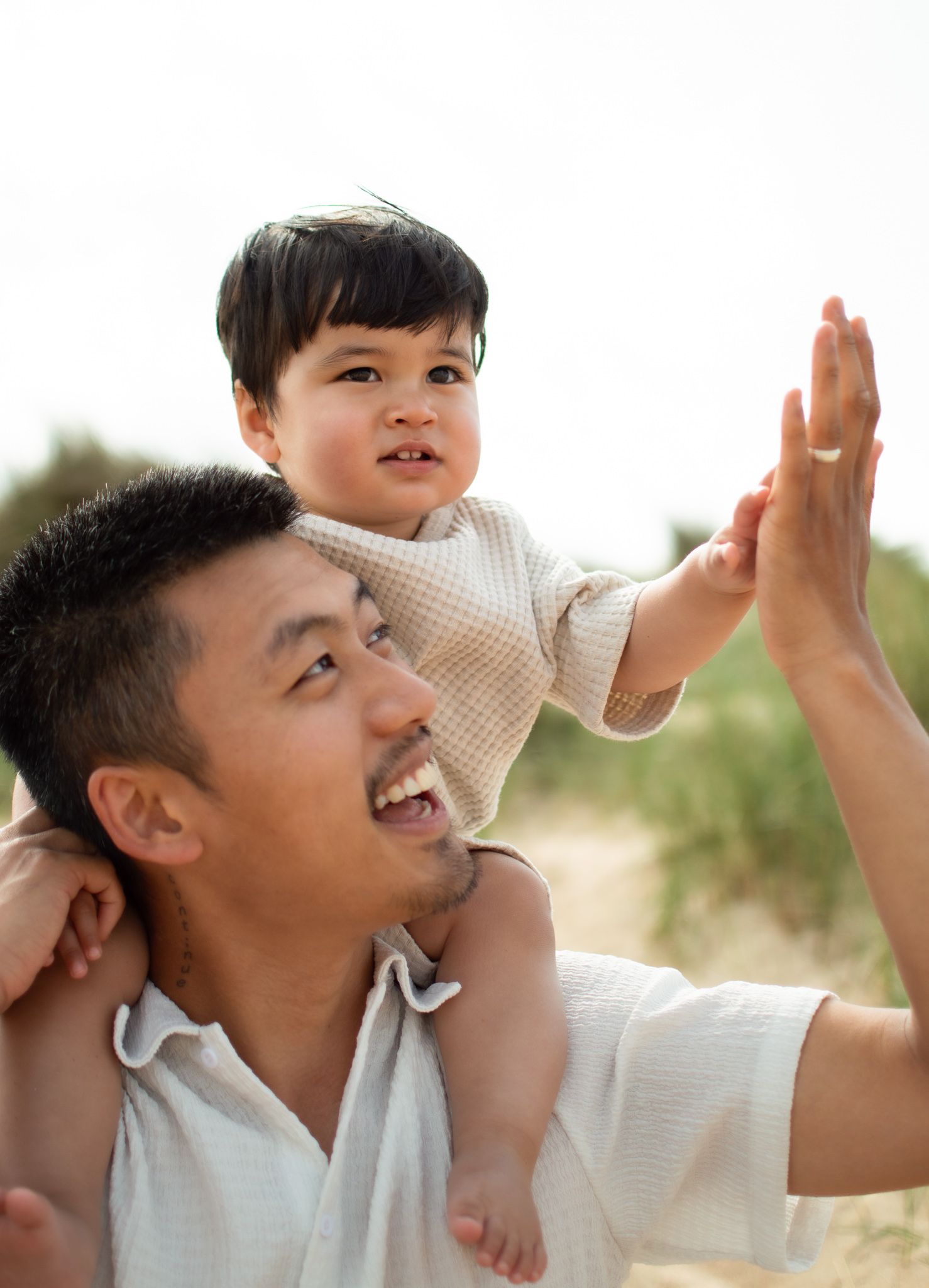 Laughing father gives toddler a shoulder ride as child reaches up for a high five at the beach.