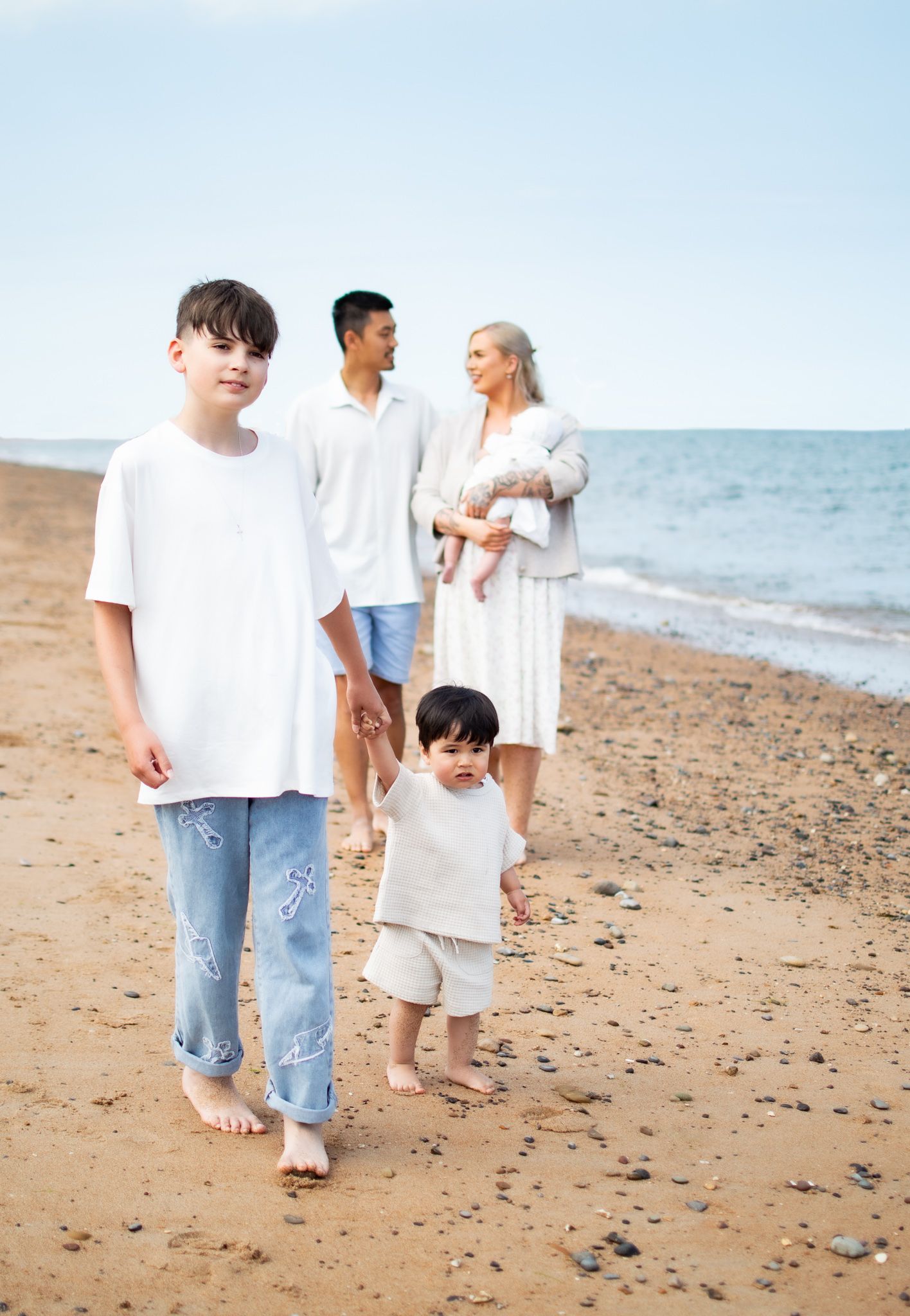 Family of five walking barefoot on a sandy beach, with two boys in front and parents holding a baby behind.