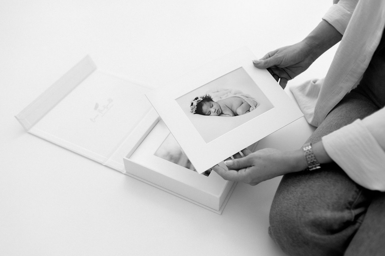 Person holding a framed newborn photo over an open white keepsake box in a black and white portrait.