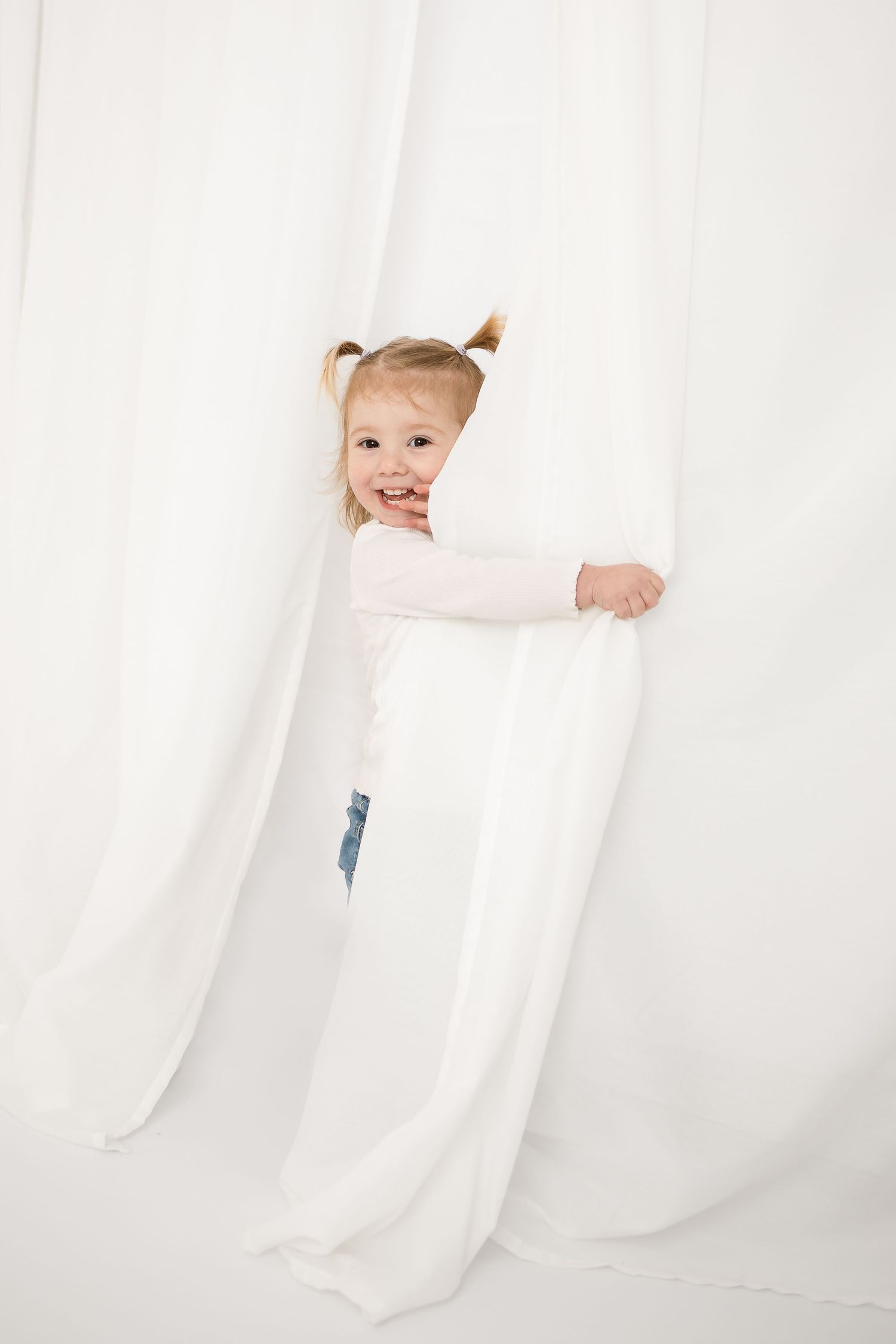 Cute toddler girl with pigtails peeking through white curtains, smiling playfully in a bright white studio setting.