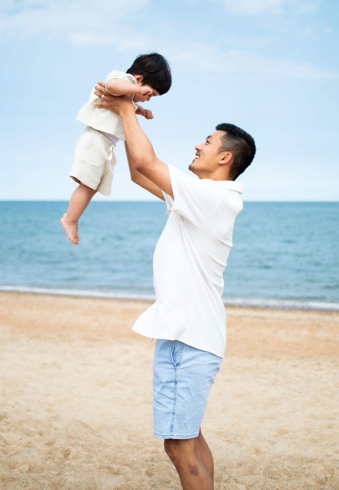 Father joyfully lifts his young child overhead on a sunny beach with blue ocean waves in the background.