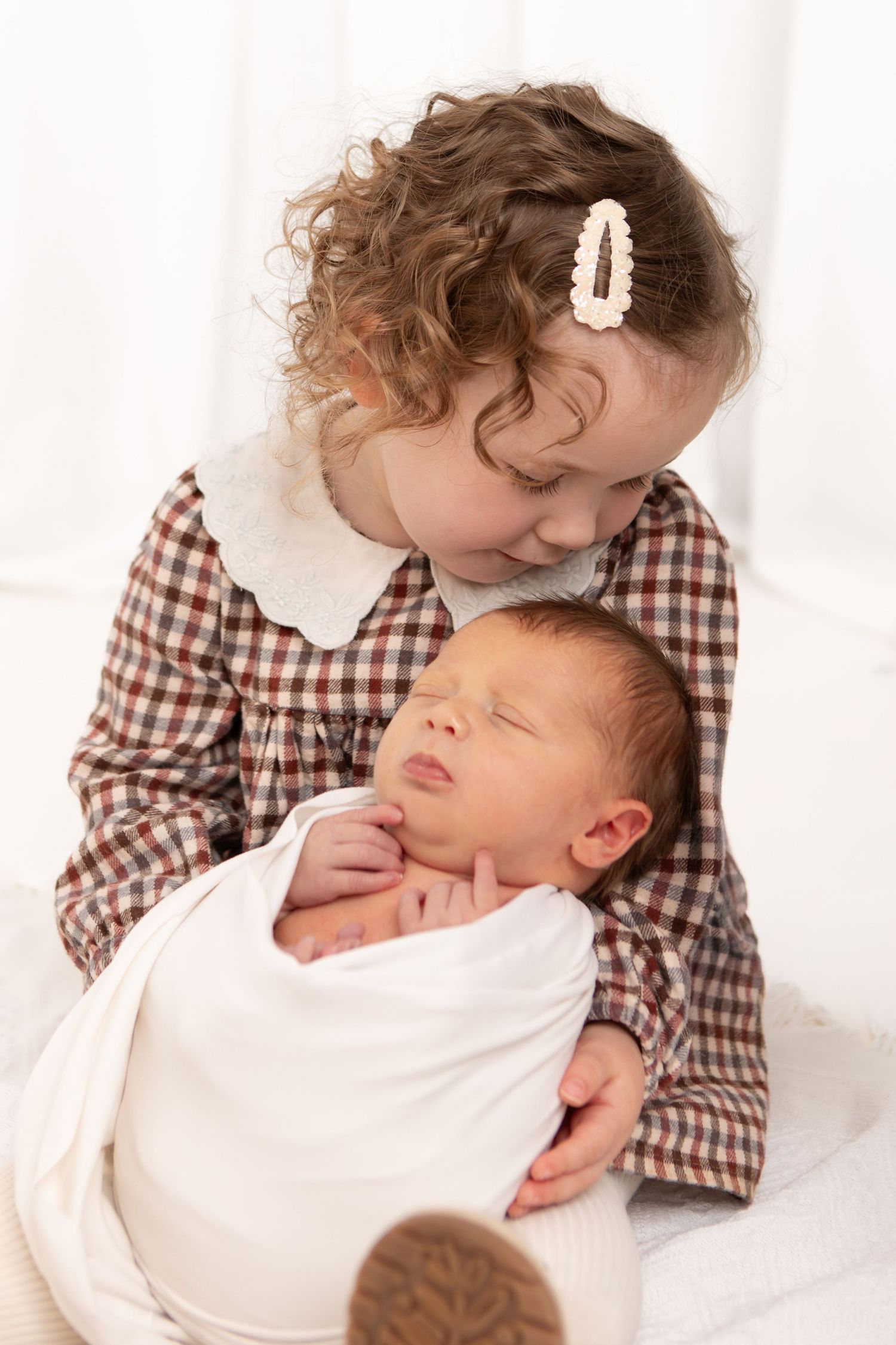 Sweet interaction between toddler in gingham dress and sleeping newborn sibling.