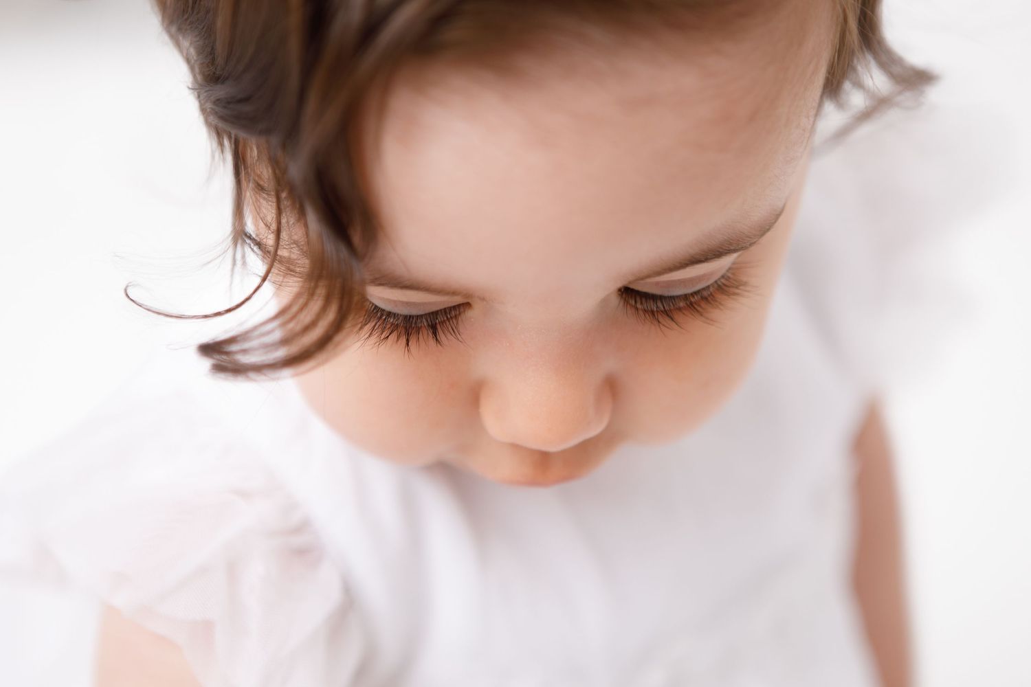 Close-up portrait of a cute baby girl with long eyelashes looking down, wearing a white dress.