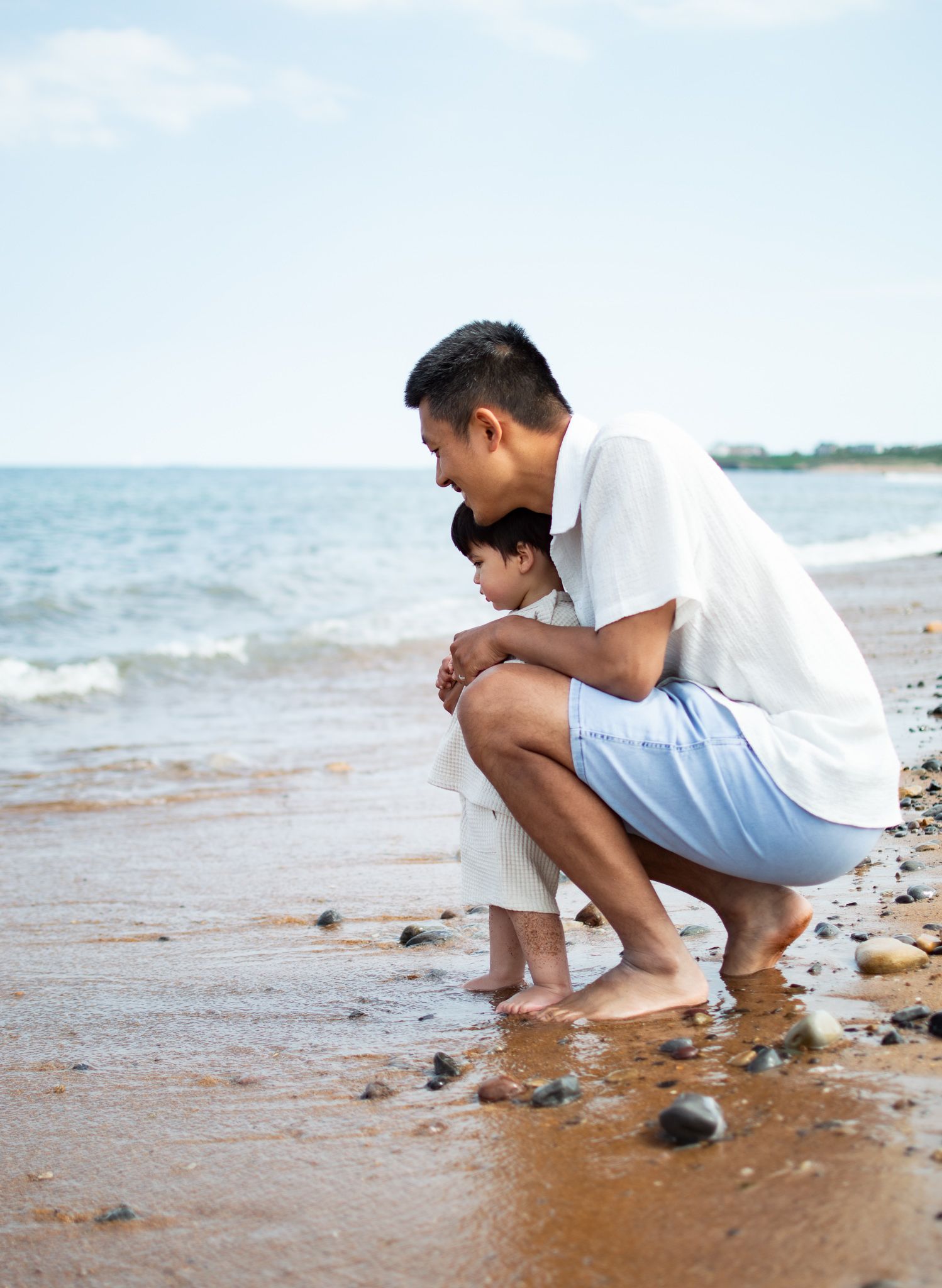 Father crouching with young son at water's edge on rocky beach, looking at stones together.