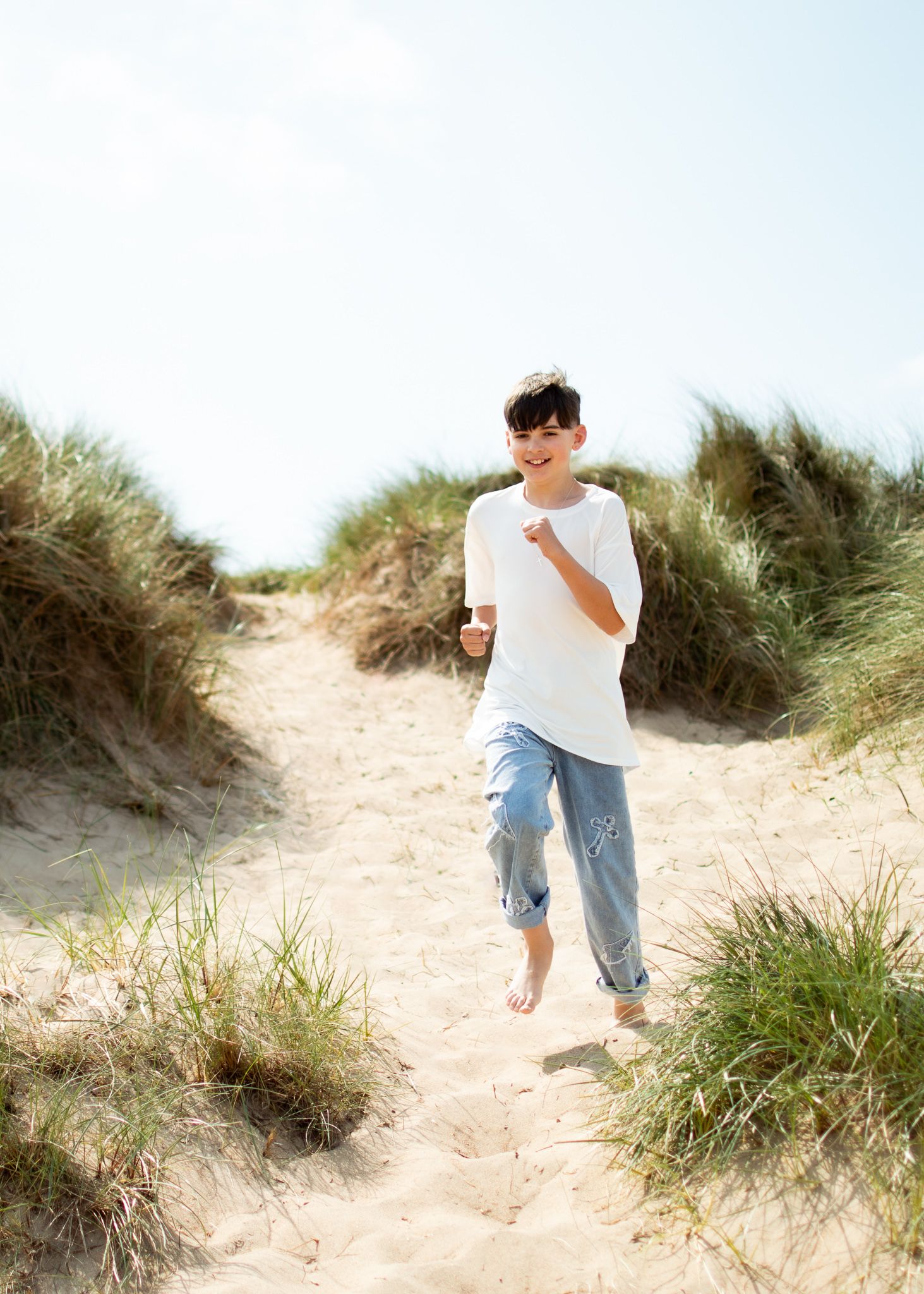 A barefoot child runs through sandy beach dunes with green grass, wearing a white tee and blue jeans.