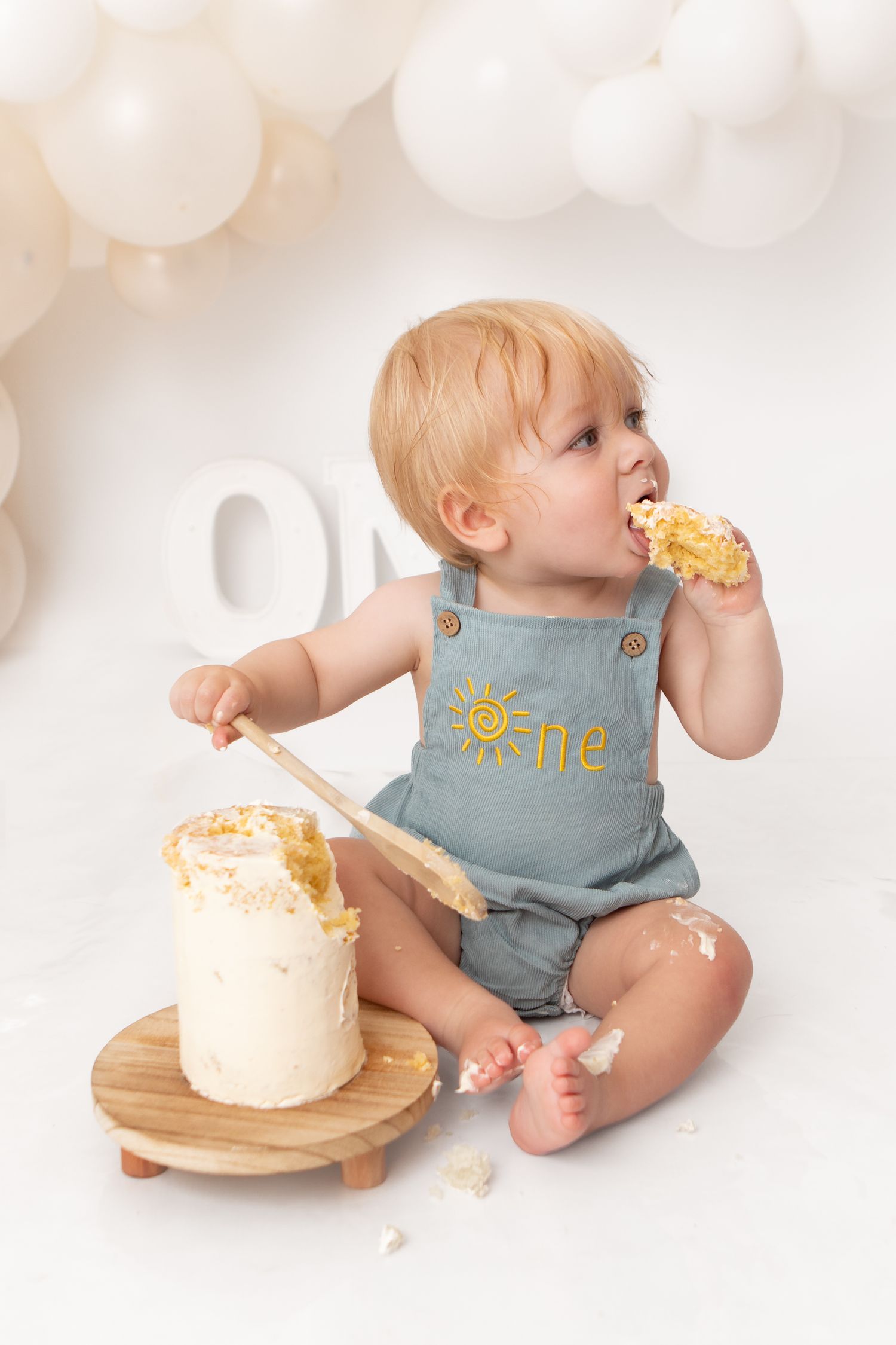 Baby in blue overalls eating smash cake at first birthday photo session with balloon backdrop.