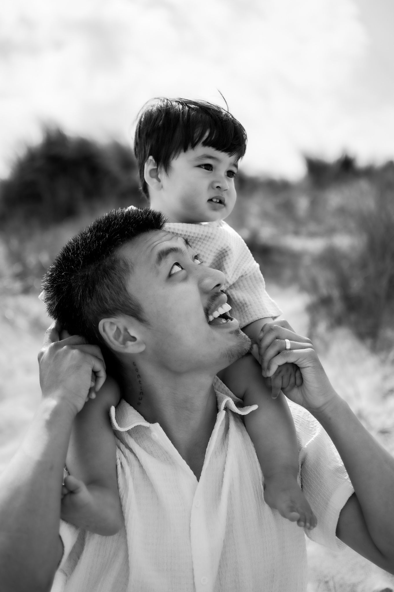 Black and white photo of father carrying toddler on shoulders at sandy beach dunes.