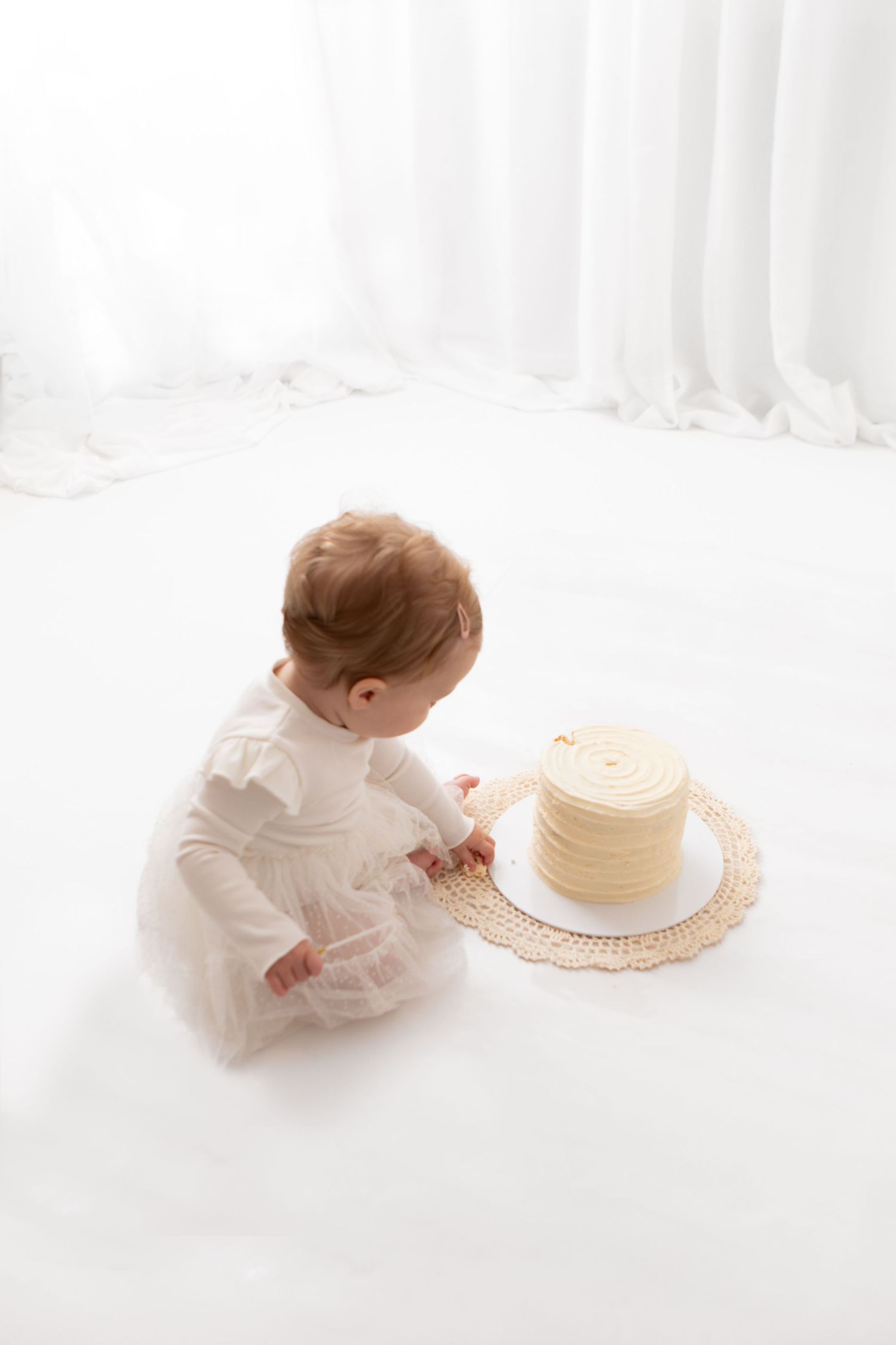 Baby in white dress sitting on floor reaching toward a vanilla birthday smash cake on a doily mat.