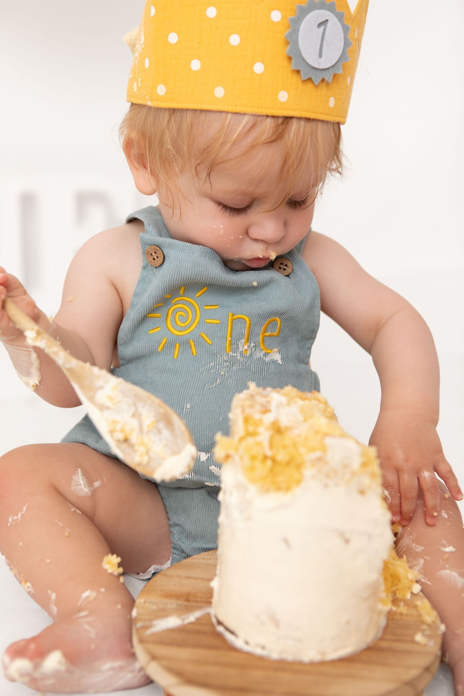 Baby in yellow crown smashes first birthday cake, wearing a sun romper with frosting everywhere.