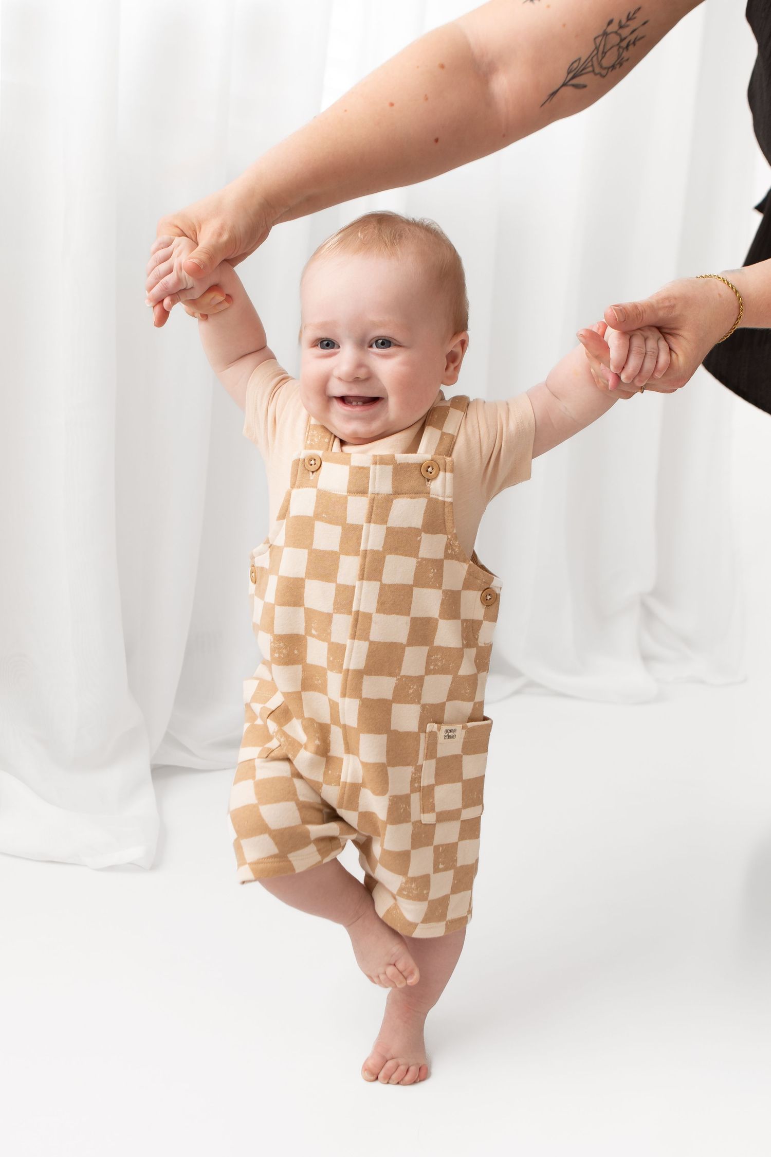 Smiling baby in tan checkered overalls stands with adult support against white backdrop.