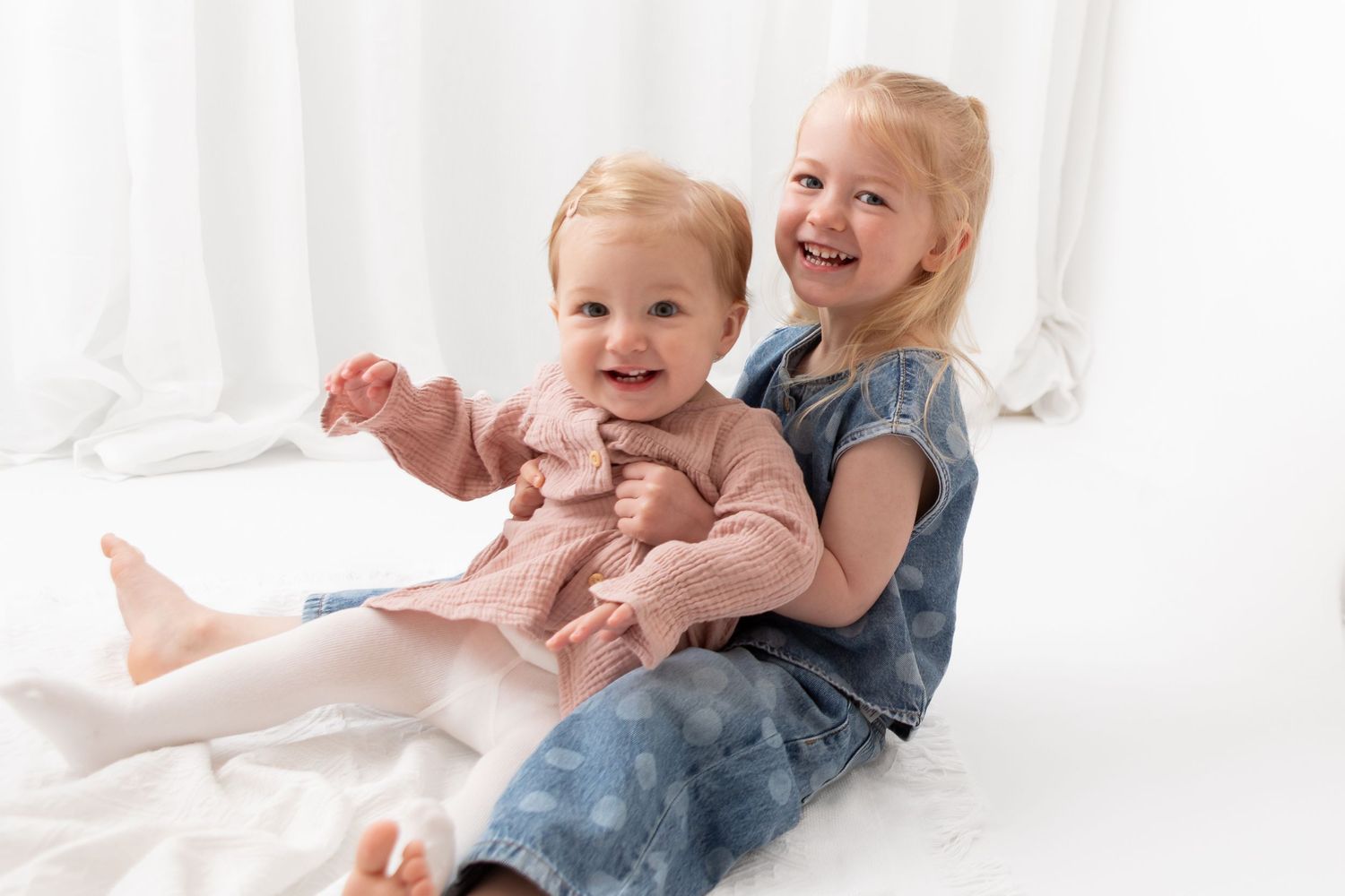 Two young sisters smiling and sitting together on a white background, wearing pink and blue dresses.