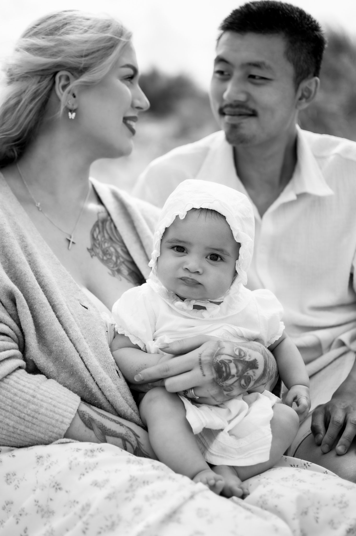Black and white portrait of smiling couple holding their baby in a white bonnet during an outdoor photoshoot.
