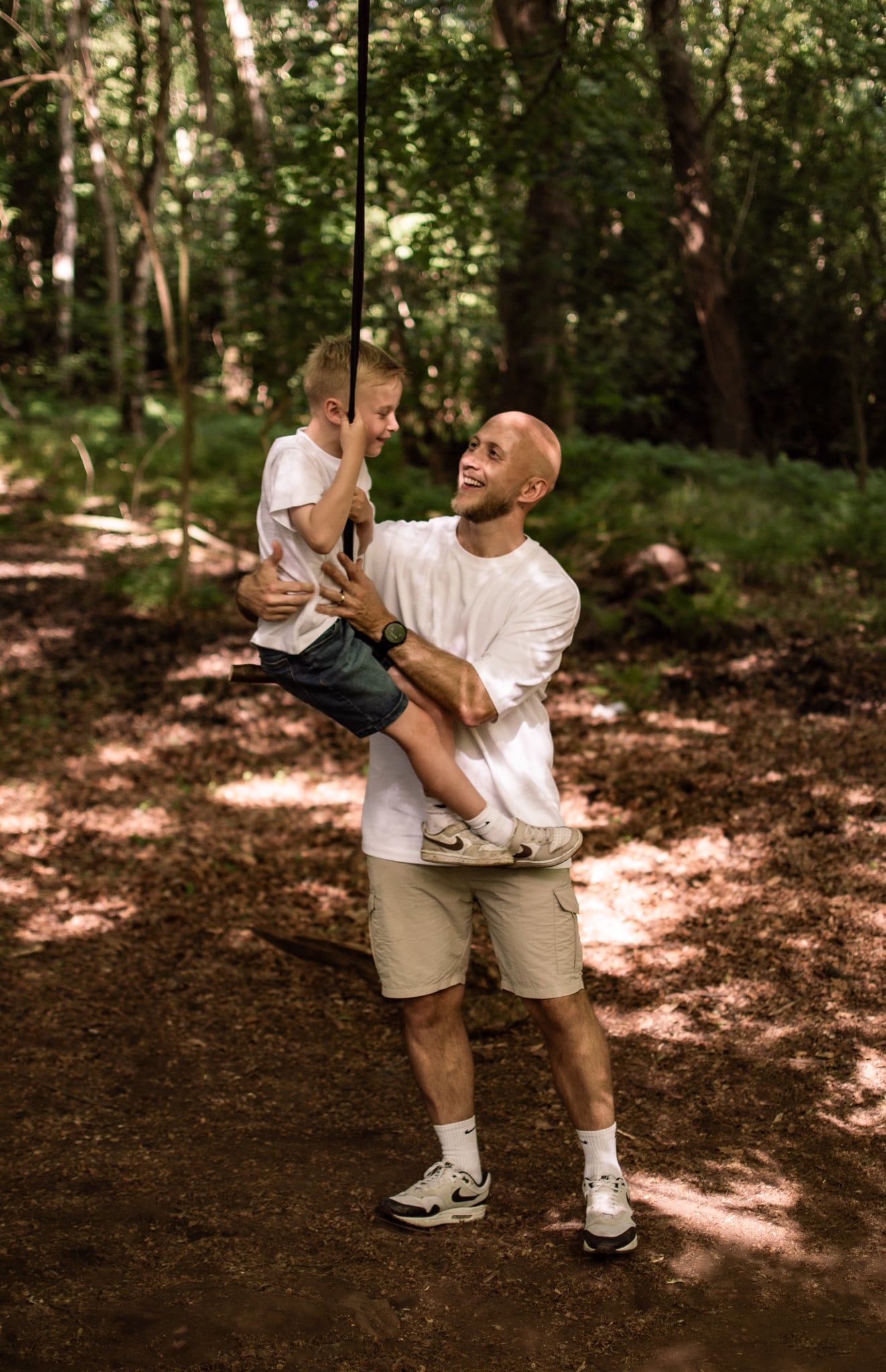 Smiling bald man holding young boy on rope swing in sunlit forest, both wearing white shirts.