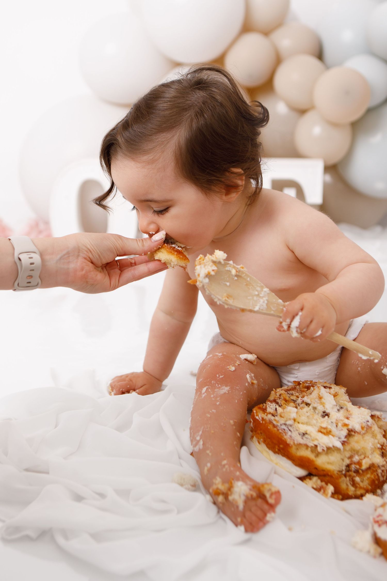 Baby enjoying first birthday cake smash, covered in frosting, sitting on white fabric with balloon backdrop.
