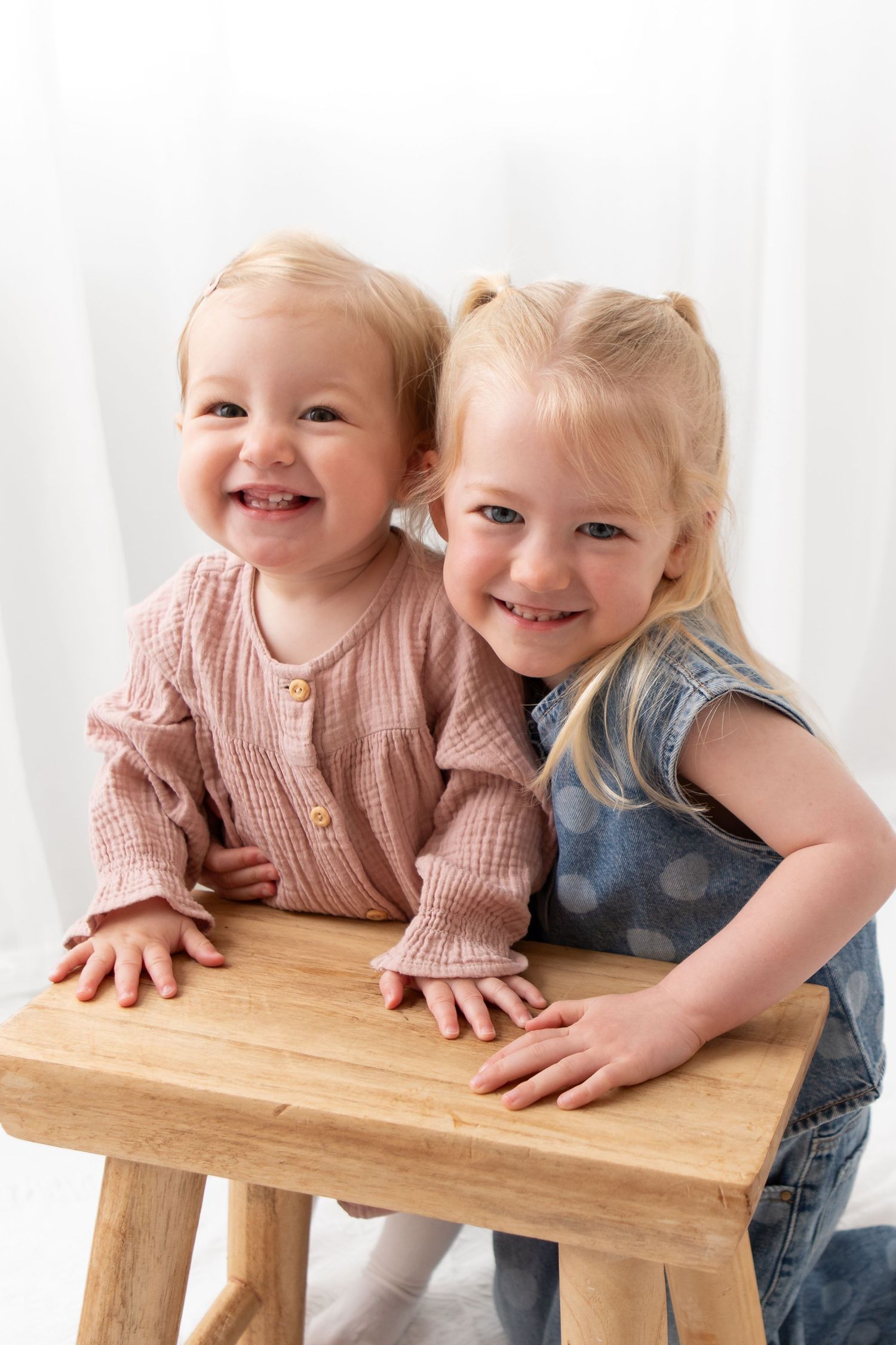 Two smiling toddler girls leaning on a wooden stool, wearing pink and denim outfits, posing for a portrait photo.