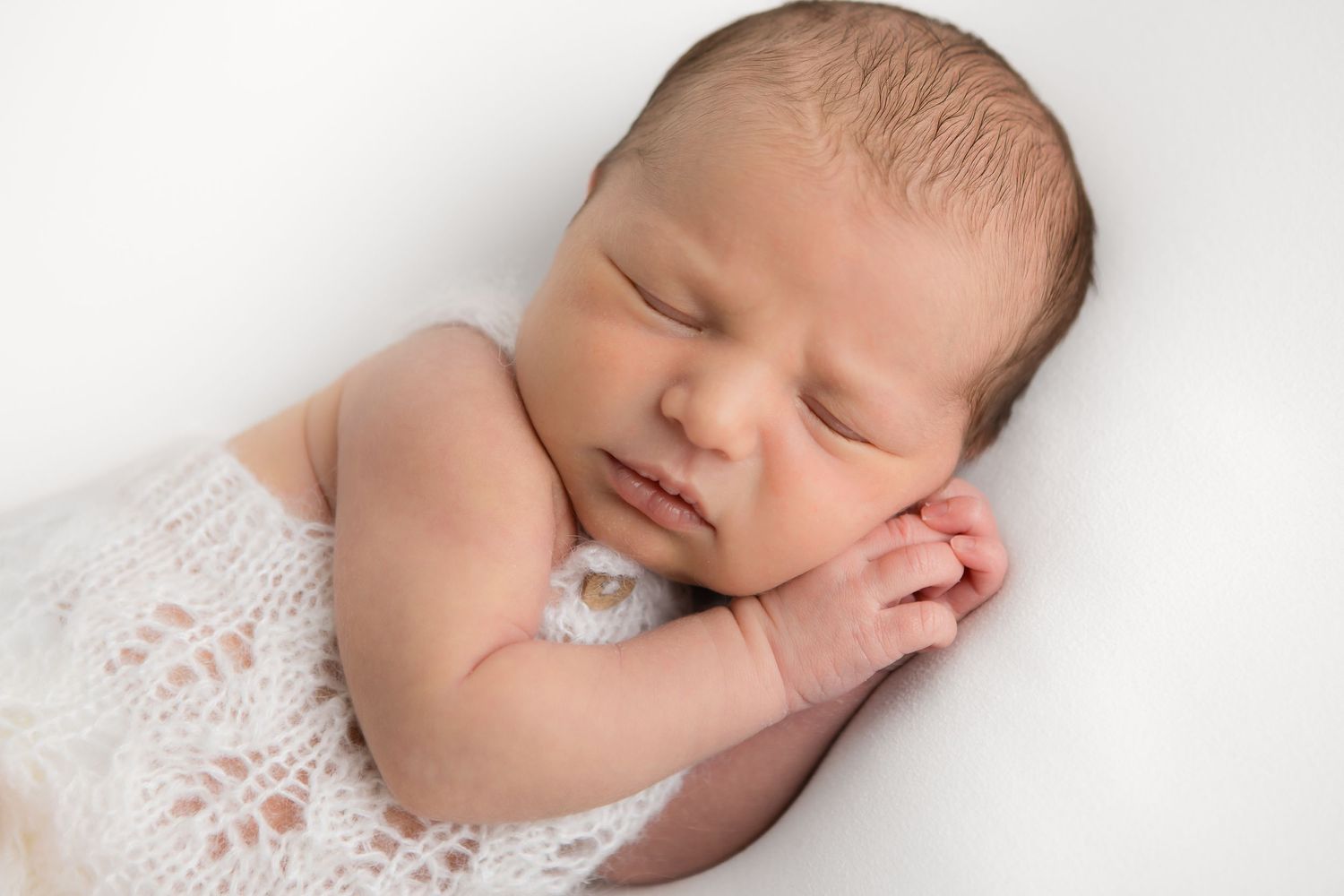 Peaceful newborn sleeping on white background with hands tucked under chin.