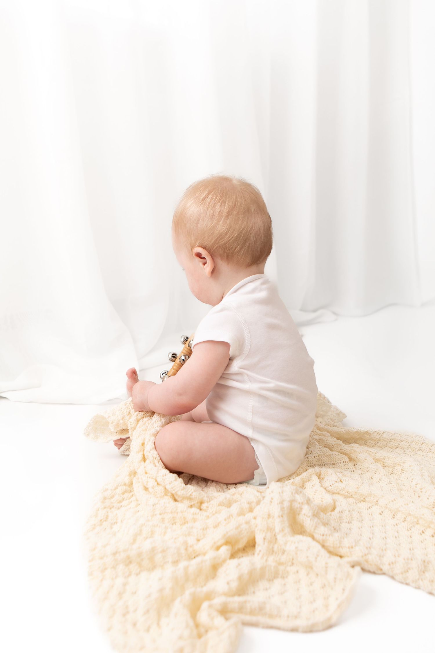 Baby sitting on a cream knit blanket in a white onesie, viewed from behind against a soft white curtain backdrop.
