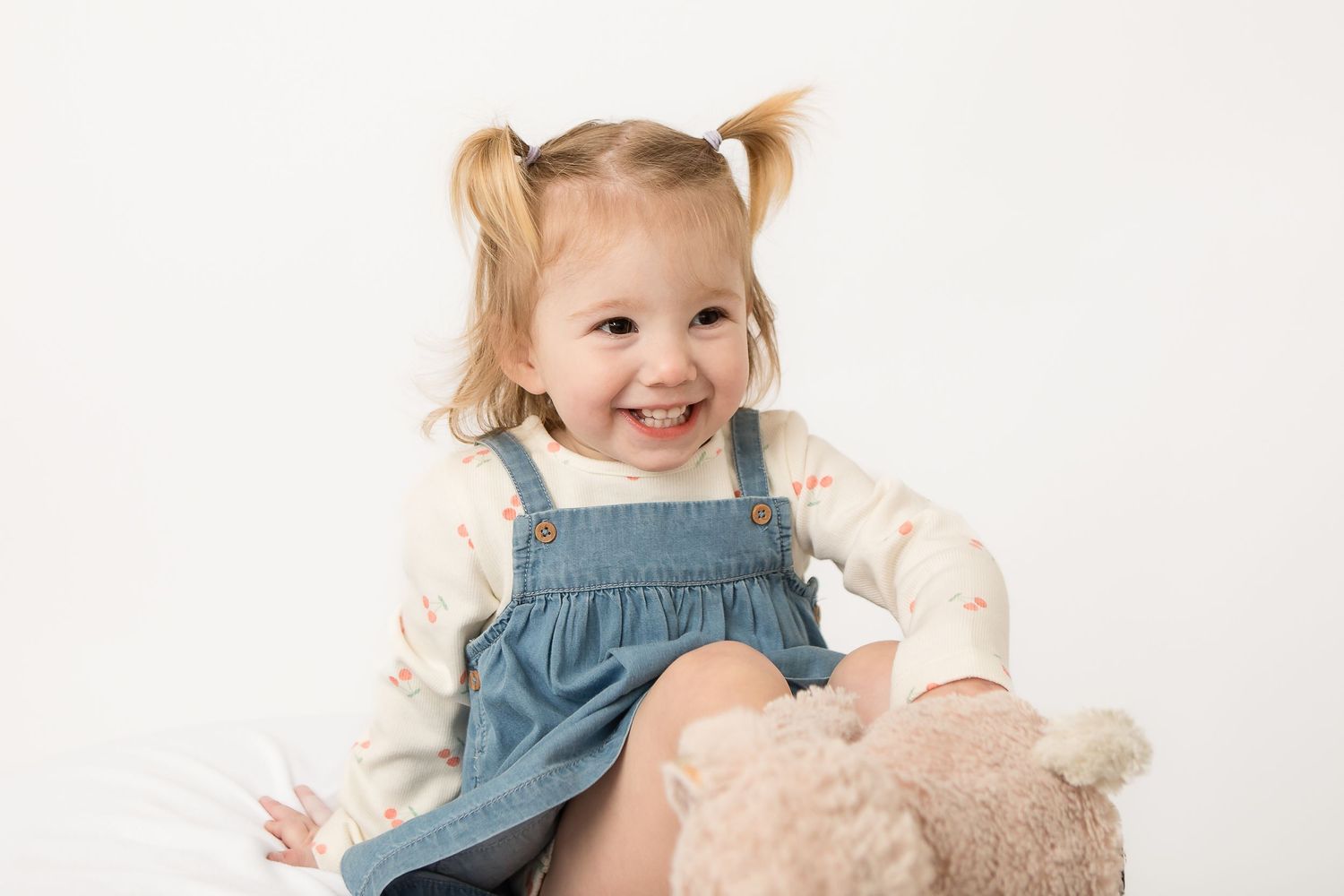 Smiling toddler girl with pigtails wearing denim overalls sitting with a teddy bear on white background.