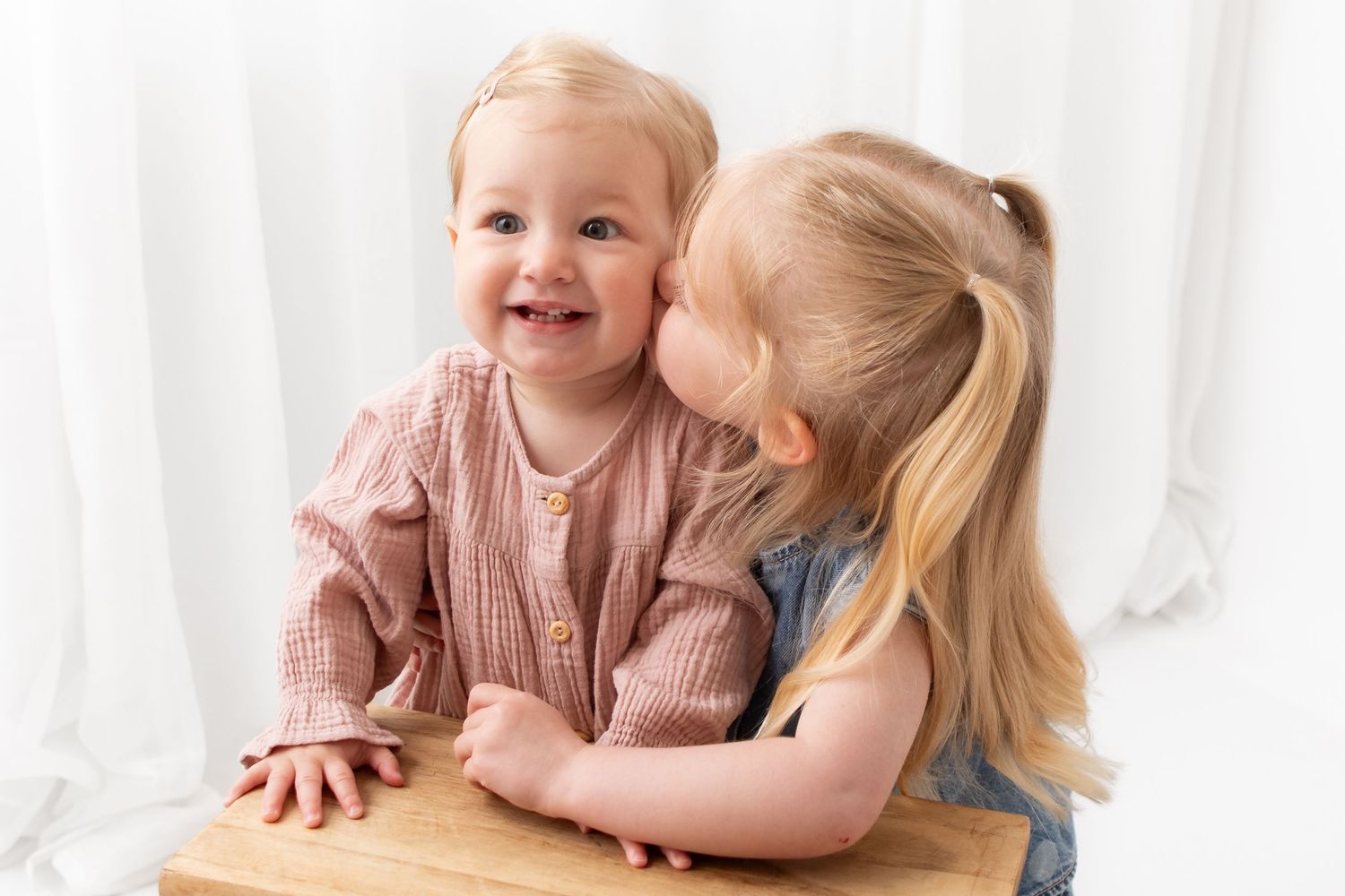 Two young blonde sisters share a sweet moment, with the older girl kissing the smiling baby on the cheek.