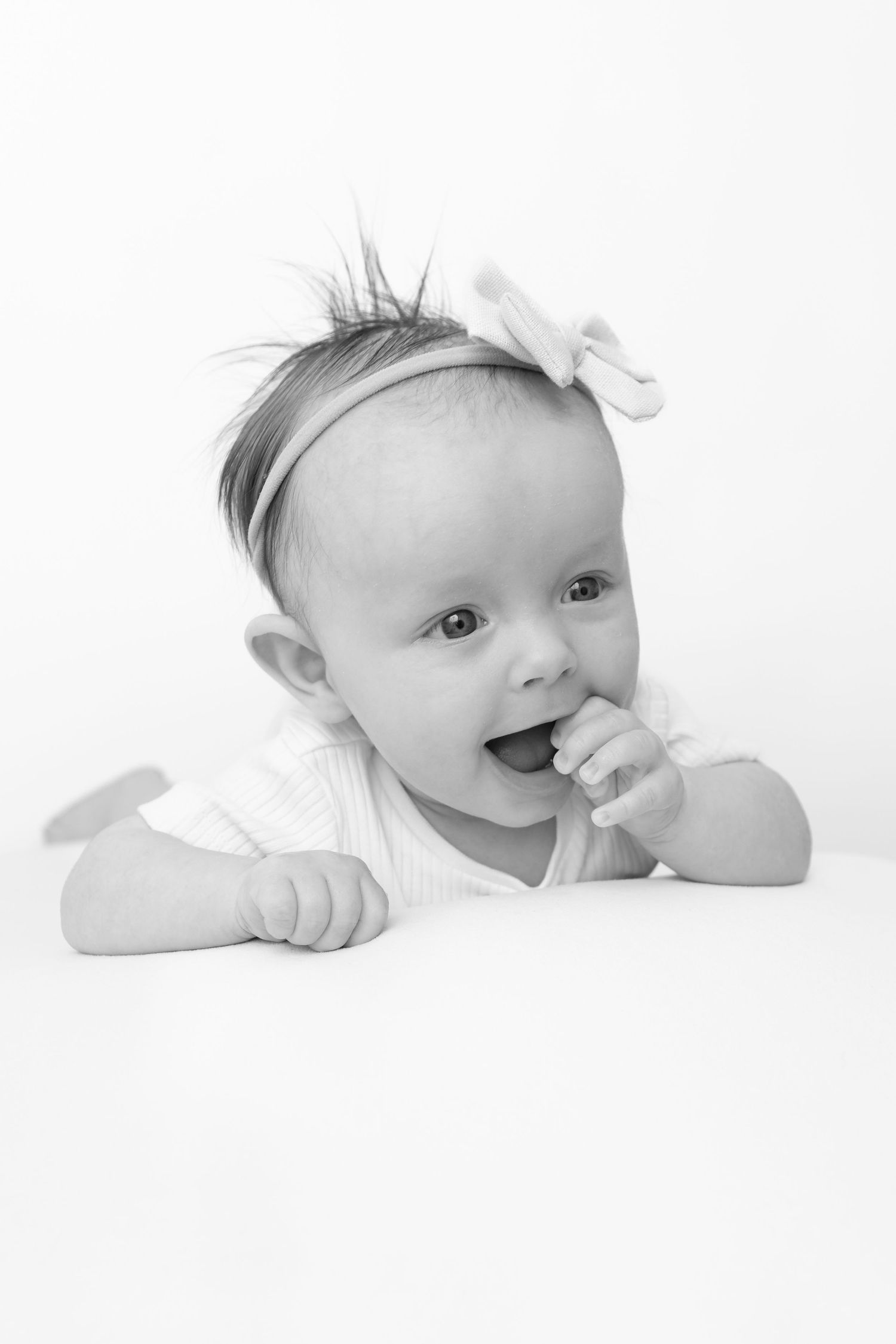 Black and white portrait of a cute baby girl with a bow headband, lying on her tummy with finger in mouth.