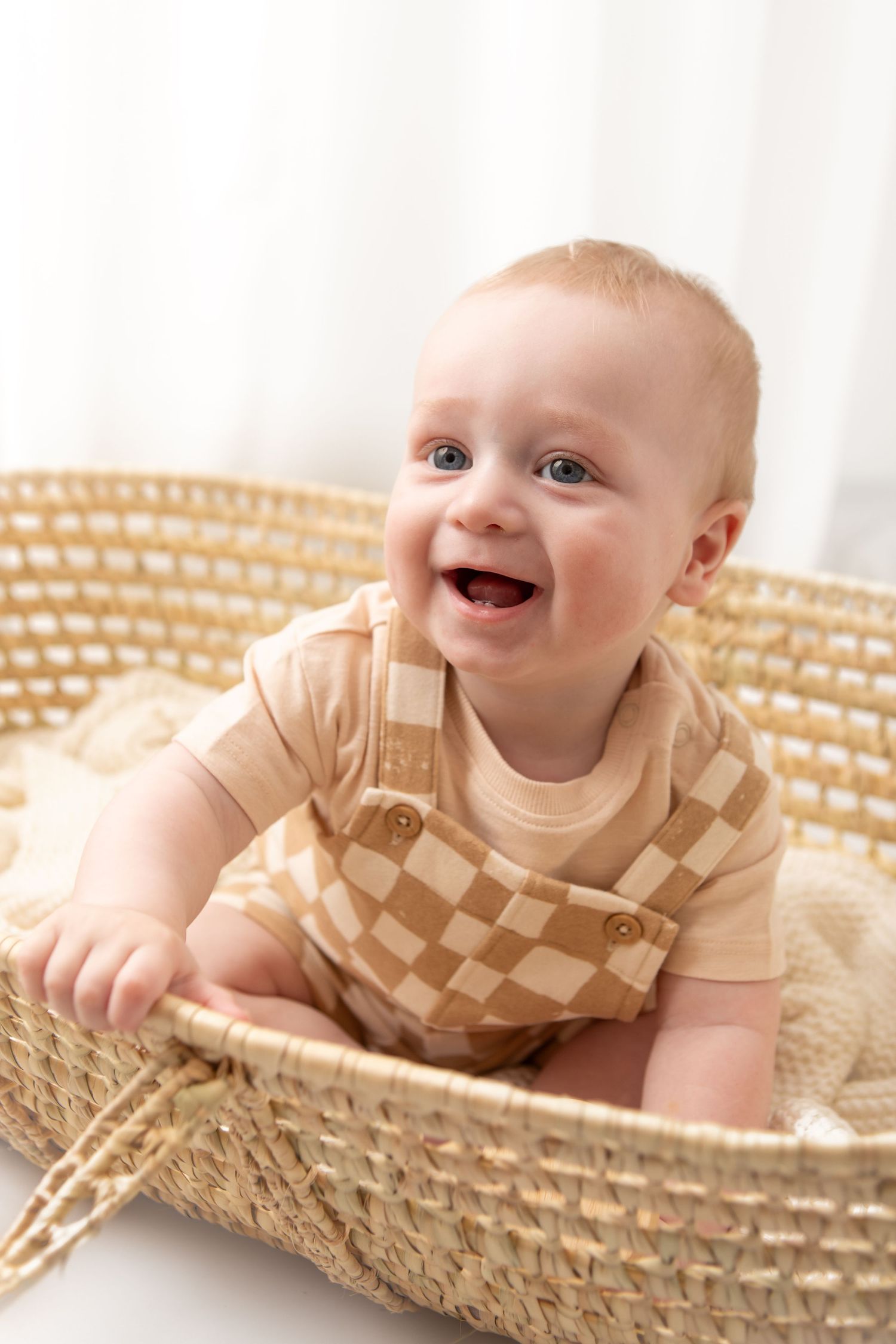 Smiling baby in a checkered beige onesie sitting in a woven basket, looking up with bright blue eyes.