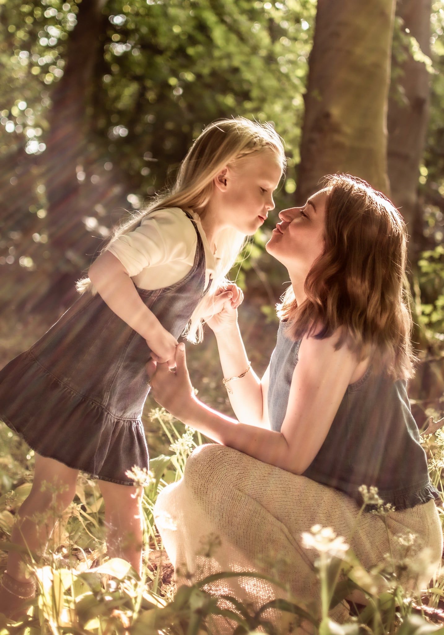 A mother and young daughter share a tender nose-to-nose moment in a sunlit forest, both wearing matching grey dresses.