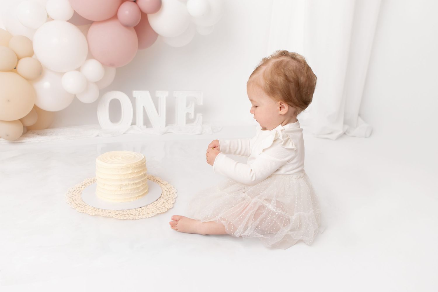 Baby girl in white dress sits beside a vanilla smash cake with ONE letters and balloon arch for first birthday.