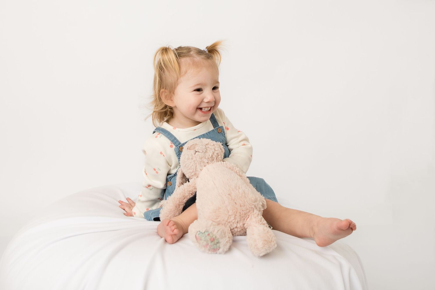 Smiling toddler girl with pigtails sitting on white surface holding a pink teddy bear, wearing denim overalls.