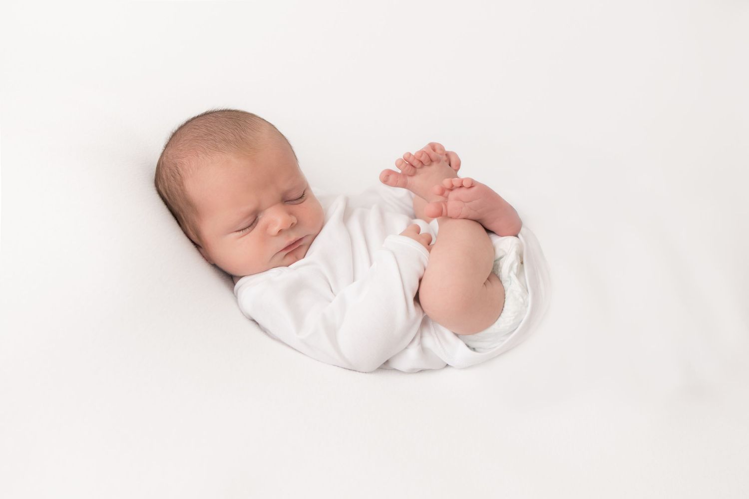 Peaceful newborn baby sleeping wrapped in white on a soft white background in a curled pose.