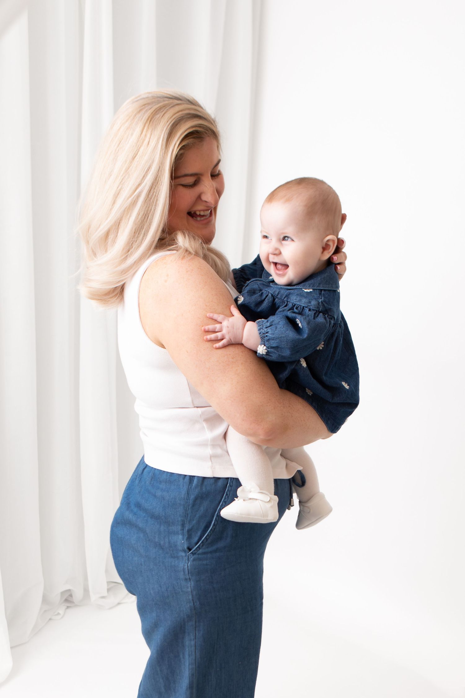 Smiling blonde woman in white tank top and jeans holding a laughing baby in a navy blue outfit against white background.