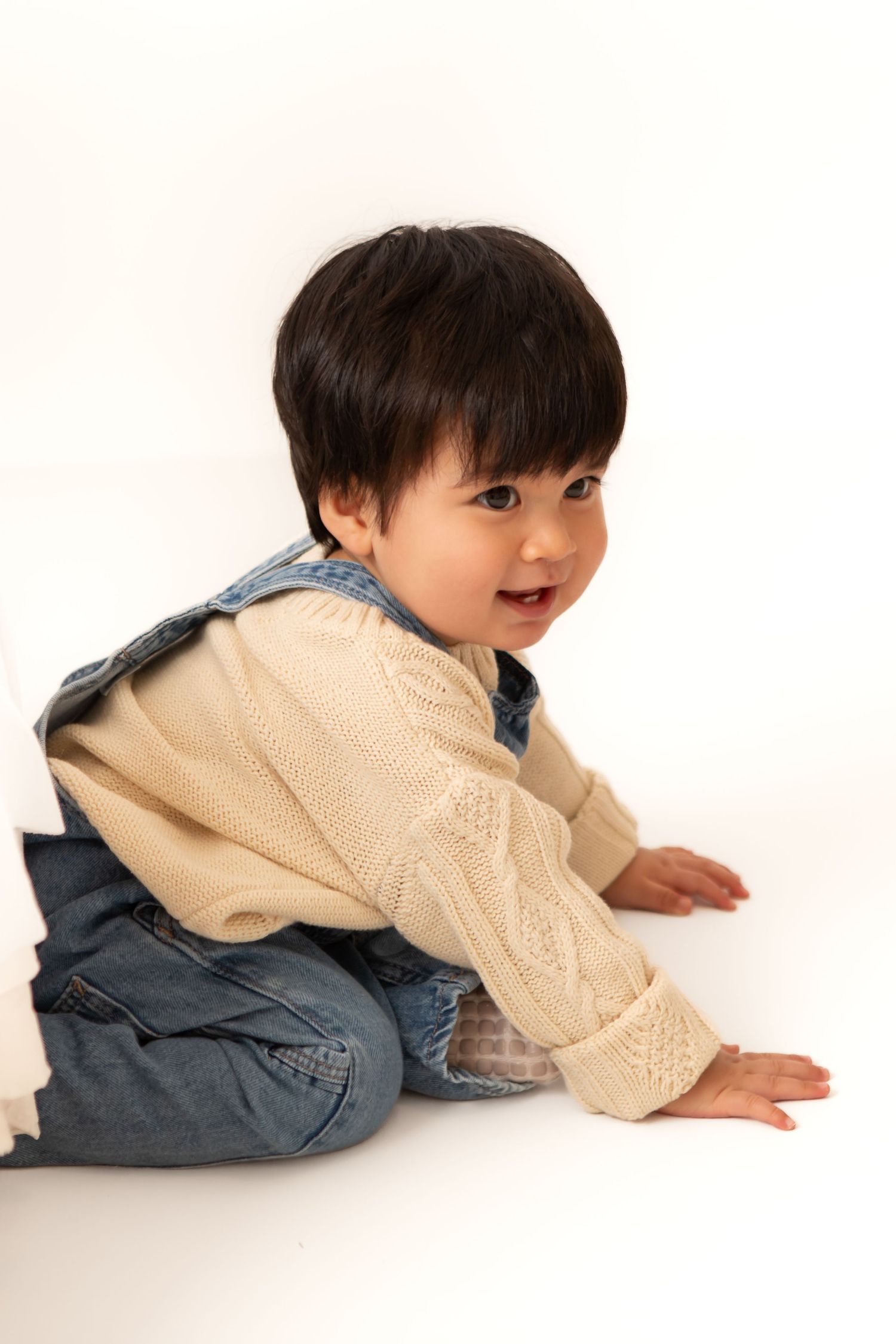 Adorable toddler in cream knit sweater and denim overalls sitting on white floor, smiling over shoulder.