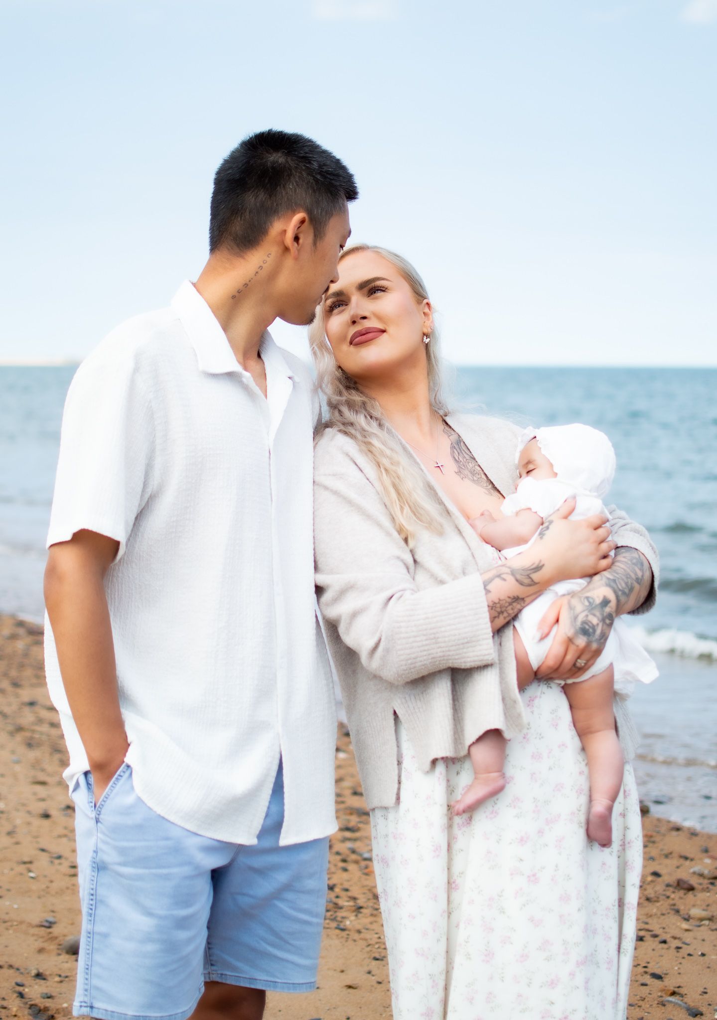 A couple standing on a beach, the woman holding a newborn baby dressed in white, sharing a tender moment.