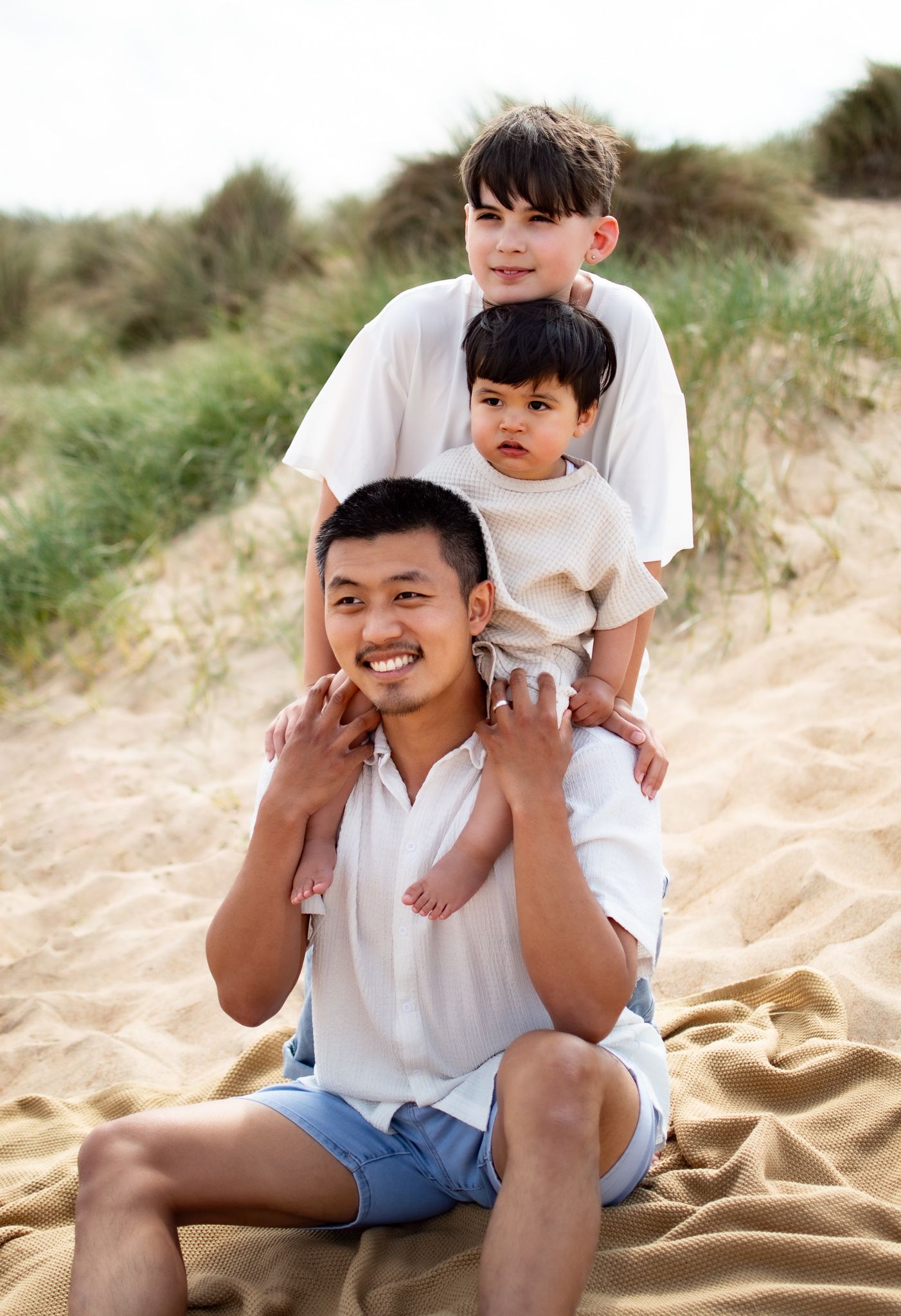 A smiling man sits on a sandy beach blanket with two young boys stacked behind him in a playful pose.