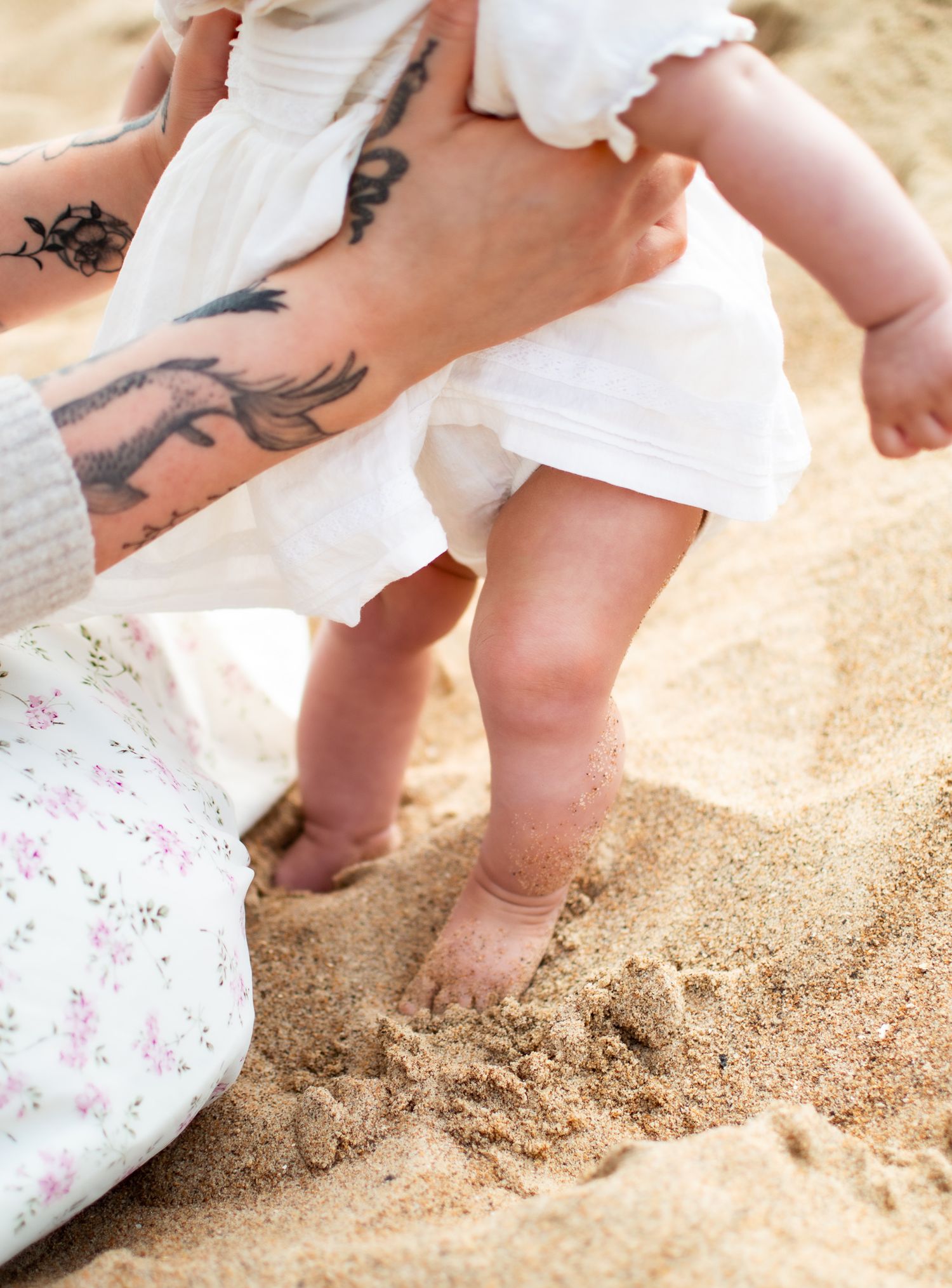 Mother with tattoos helping baby girl stand on sandy beach.