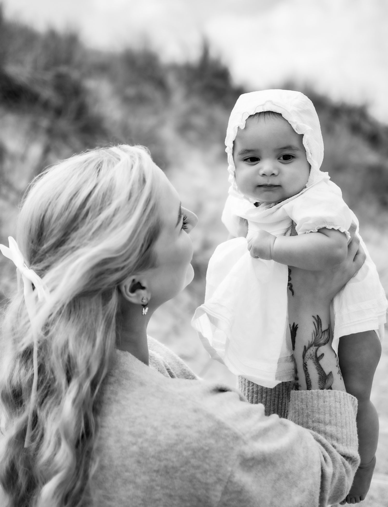 Black and white photo of a smiling baby in a bonnet being held up by a blonde woman on a beach.