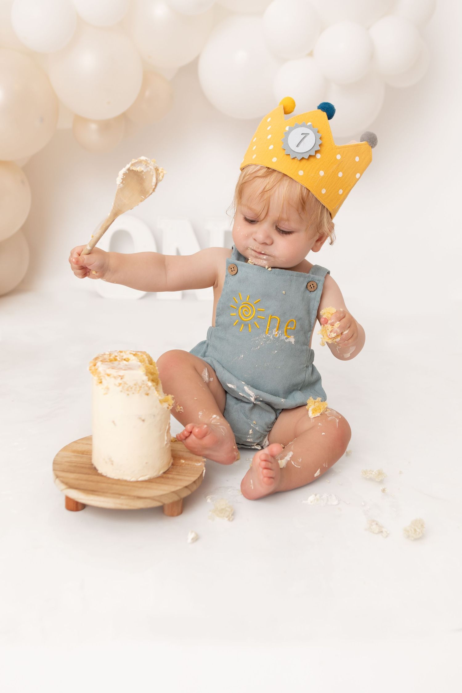Baby in yellow crown smashes first birthday cake with wooden spoon, wearing blue 