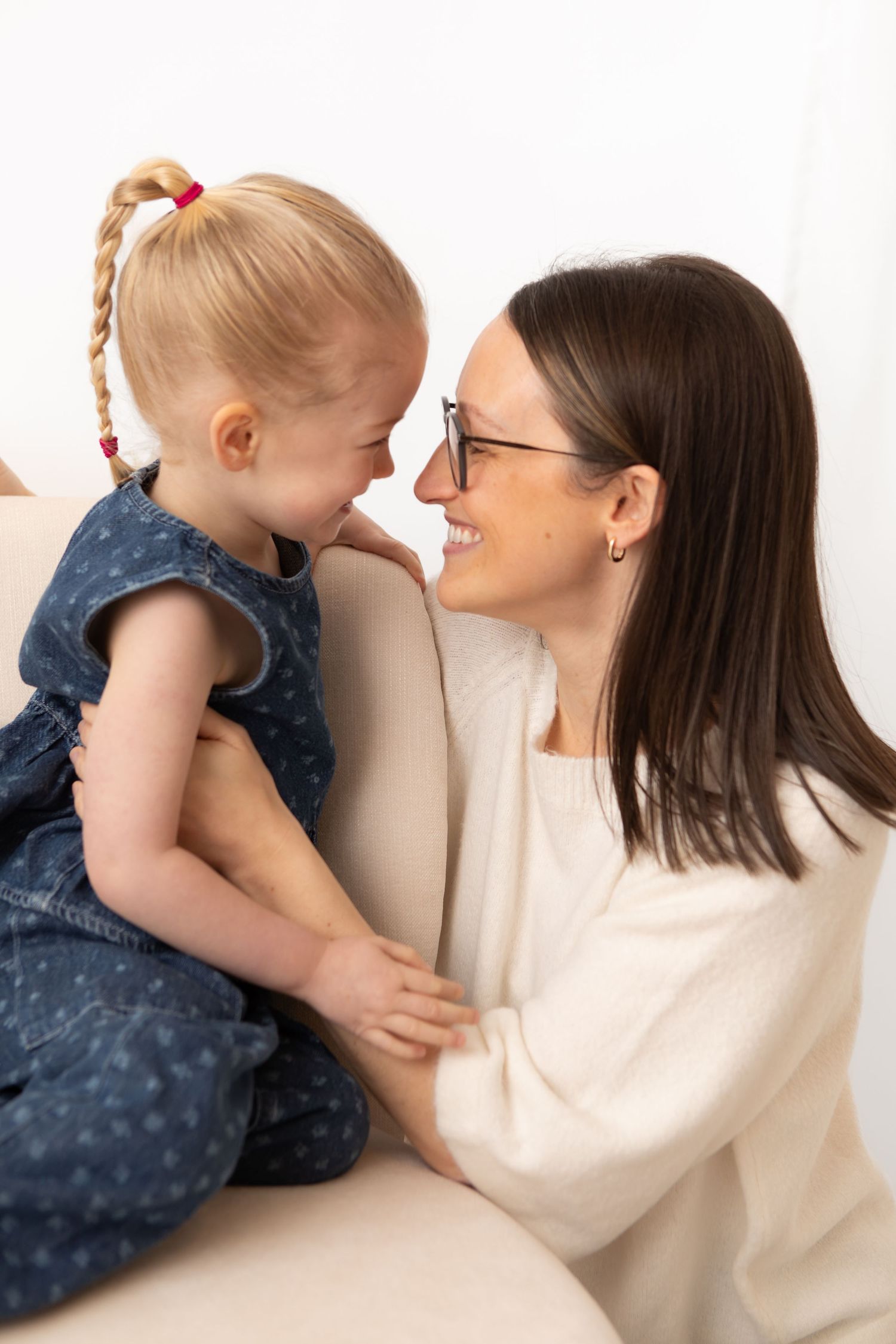 A smiling mother with glasses and dark hair shares a joyful nose-to-nose moment with her young blonde daughter on a sofa.
