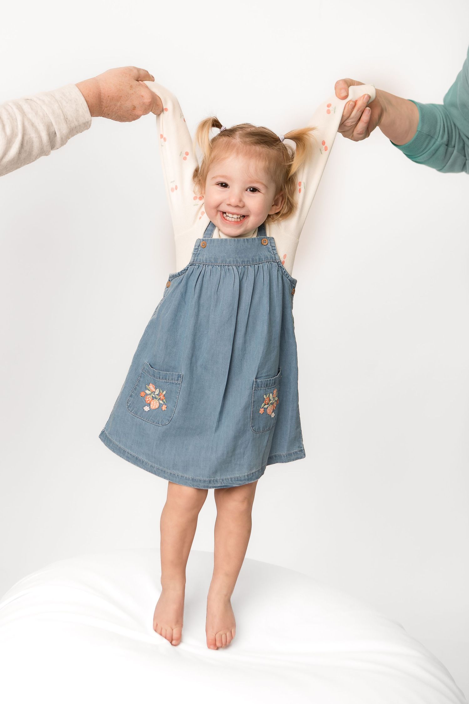 Smiling toddler girl in denim dress with floral pockets, arms raised by adults, standing barefoot on white background.