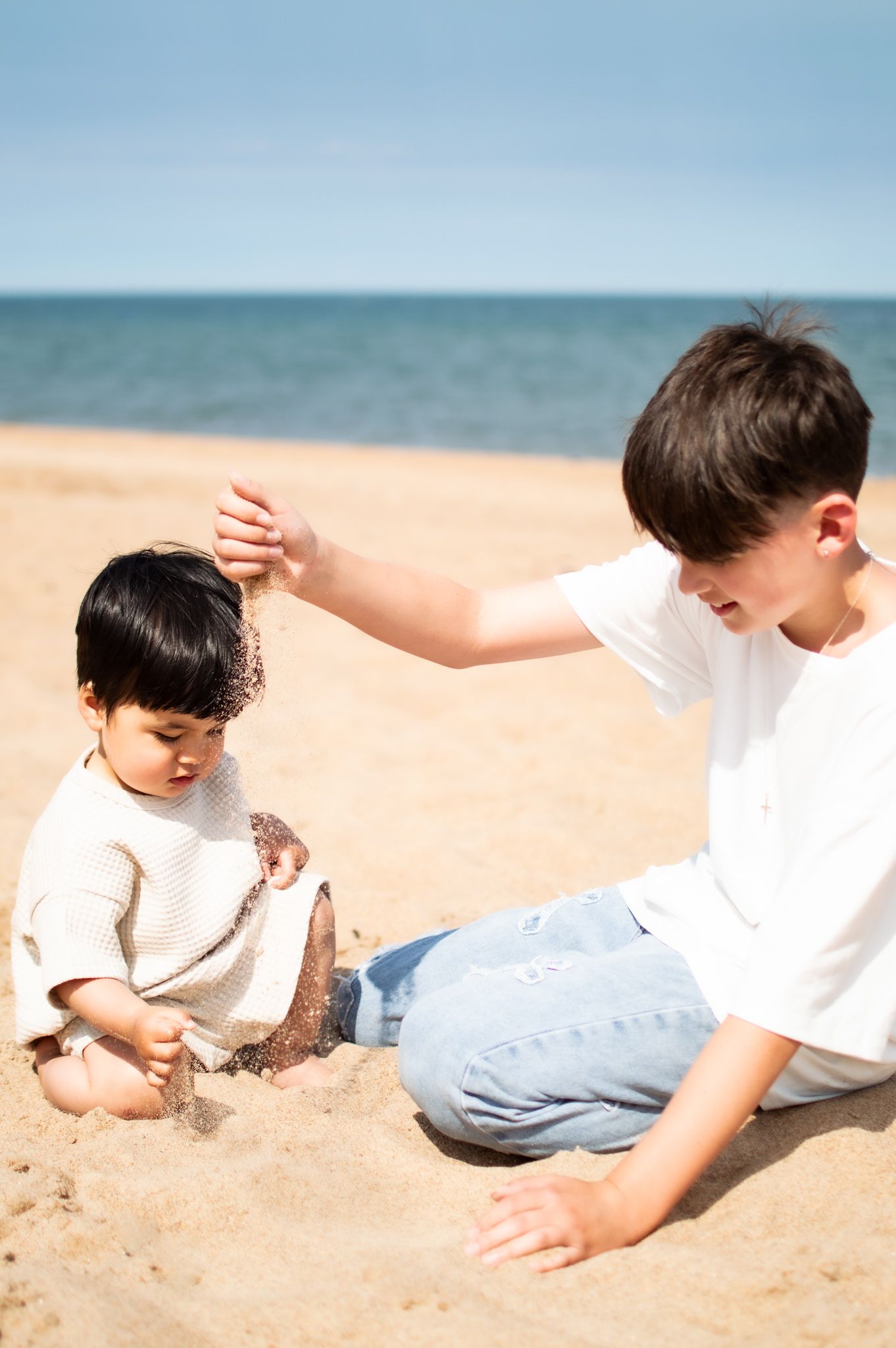Two young boys playing with sand on a sunny beach, with the ocean visible in the background.