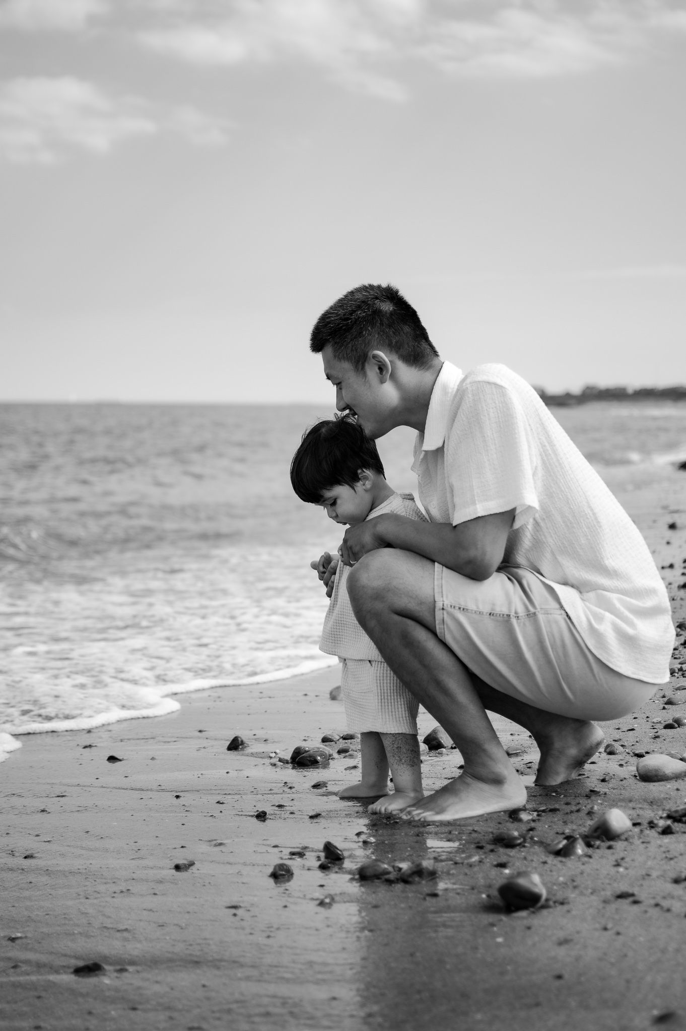 Black and white photo of a father crouching with his young child on a rocky beach, sharing a tender moment by the ocean.