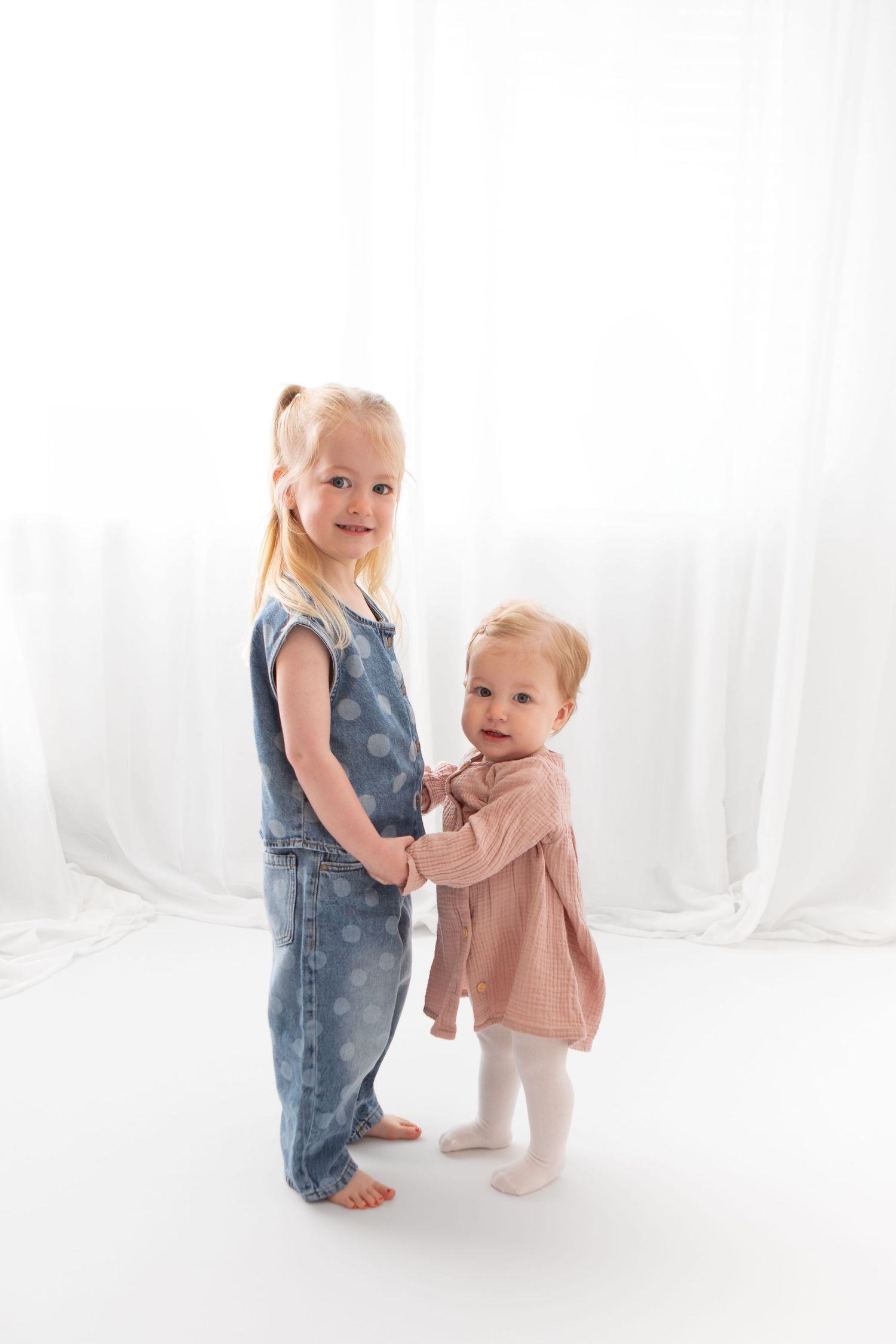 Two young sisters holding hands, older girl in denim overalls and toddler in pink dress, posing together against white backdrop.