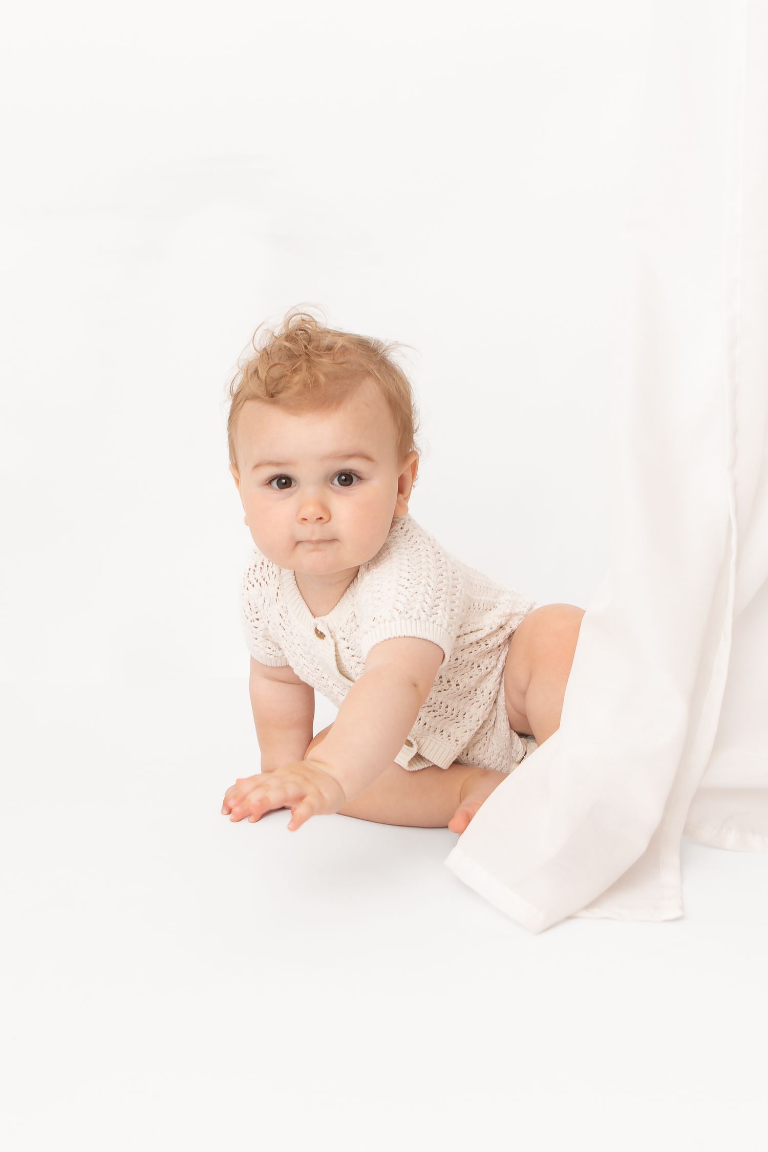 A cute baby in a cream knit romper crawls on a white background, looking curiously at the camera.