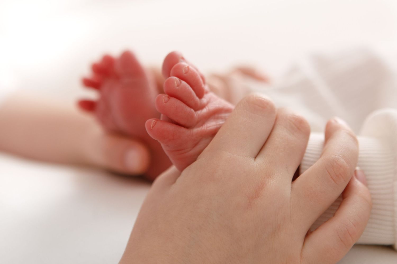 Close-up view of tiny newborn baby feet and hands being gently held against a soft white background.