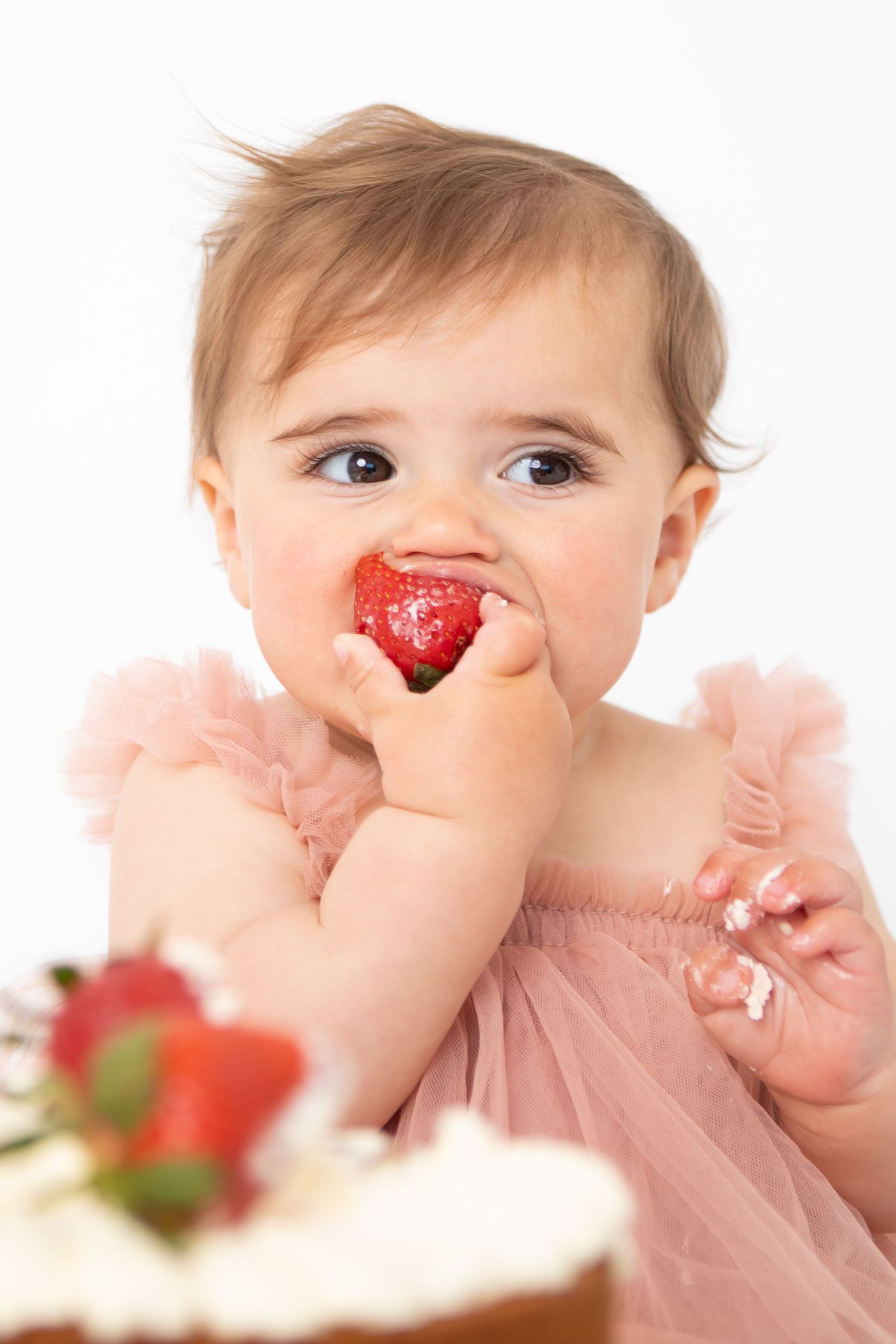Baby girl in pink tutu dress eating a strawberry in front of a birthday cake on white background.
