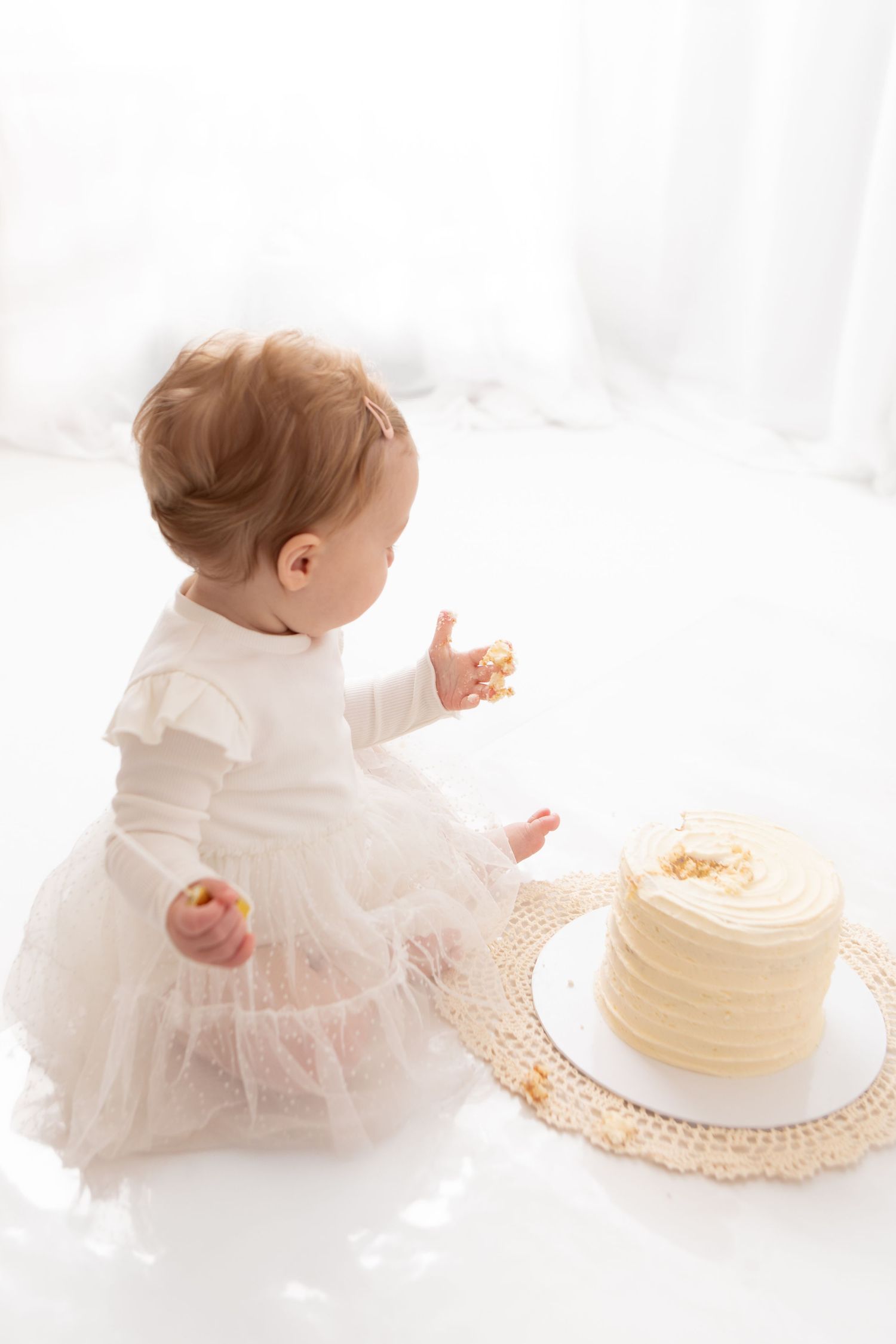 Baby girl in white tutu dress touching first birthday smash cake on white background.