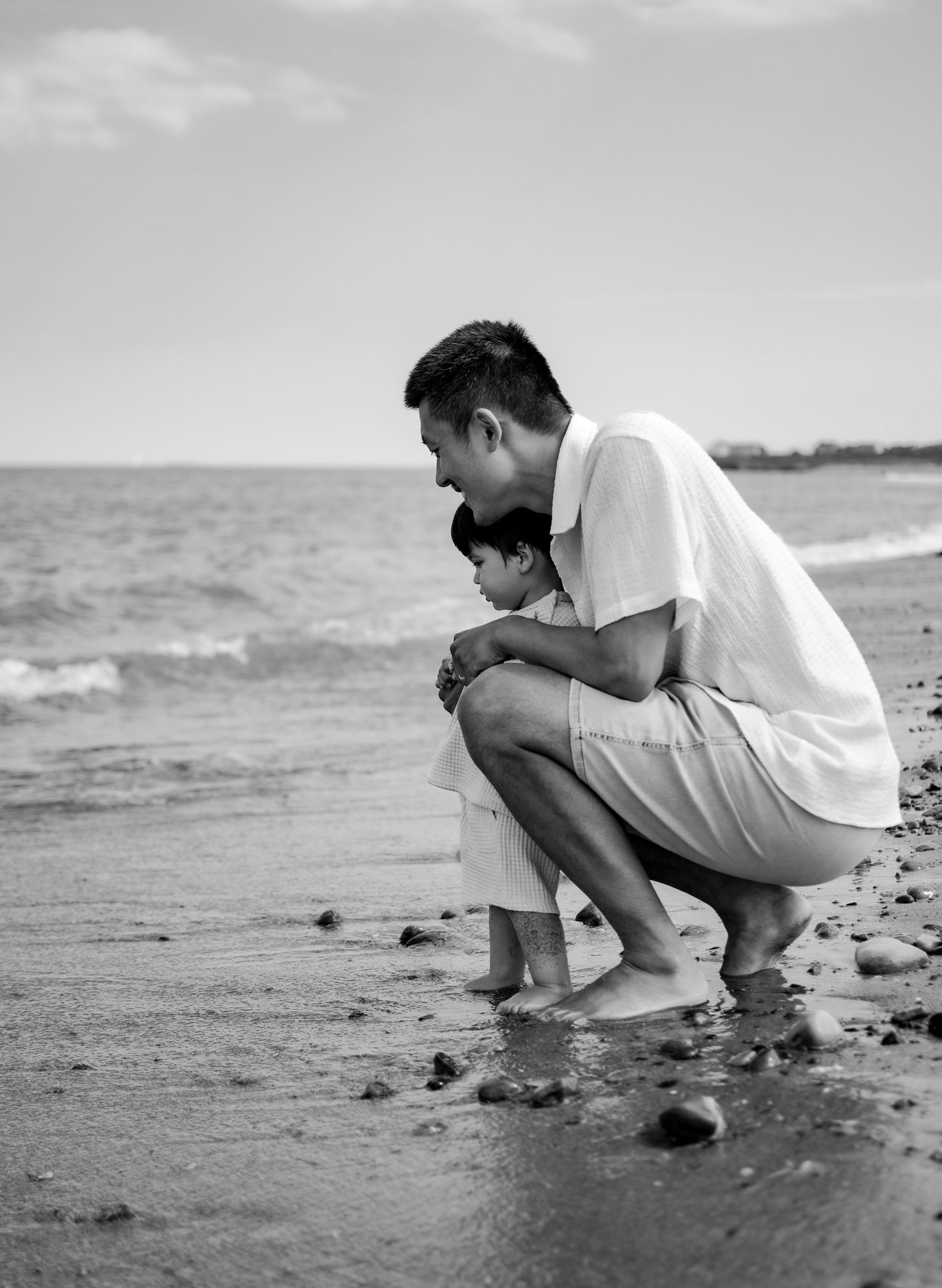 Father and young child crouch together on a rocky beach shoreline in a tender black and white photo.