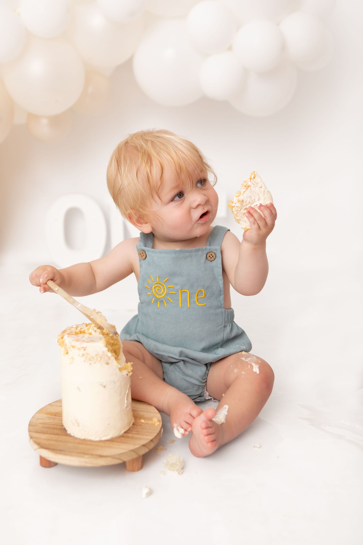 Toddler in blue overalls enjoys first birthday cake smash with white balloons and scattered frosting.