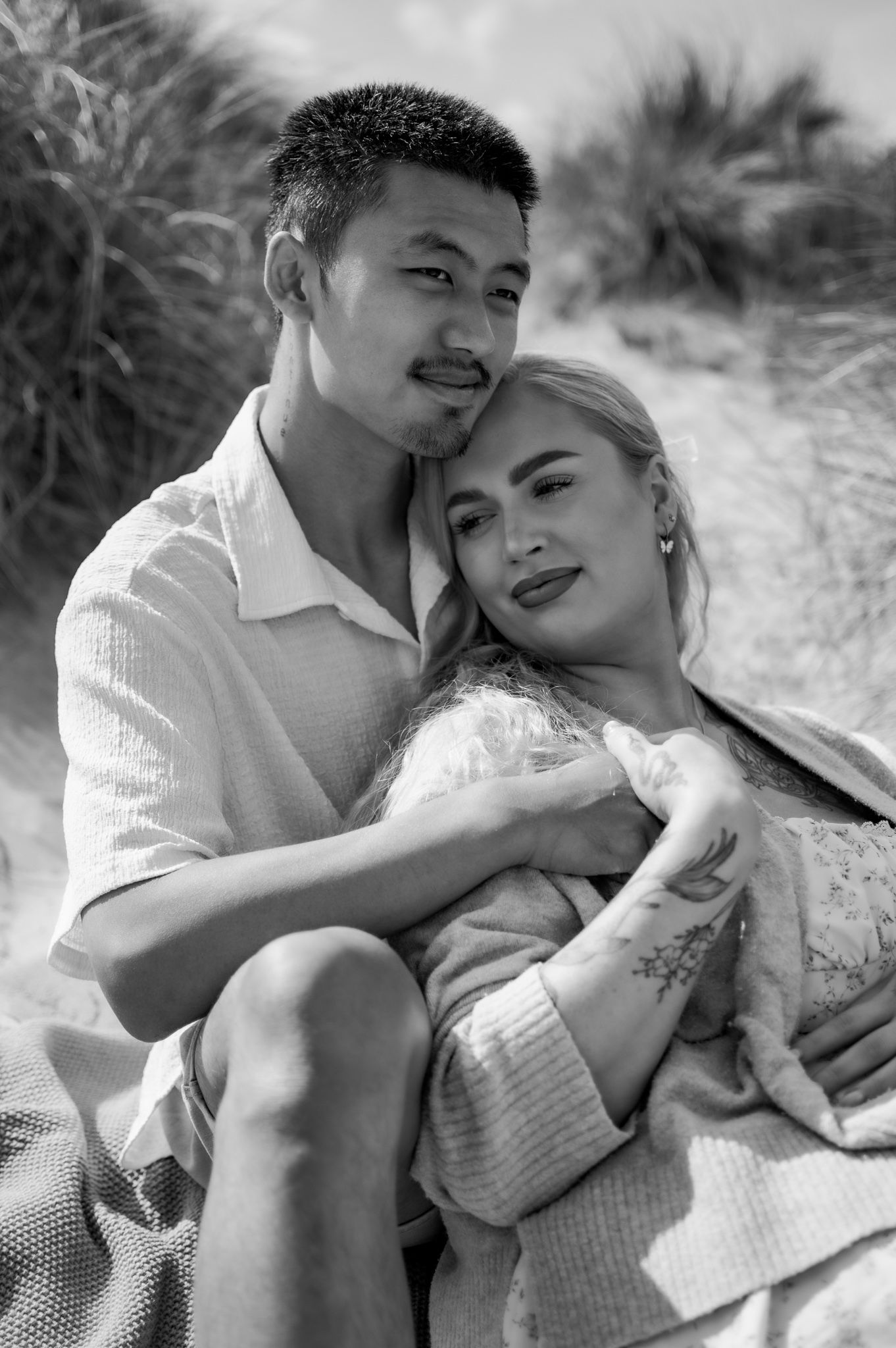 Black and white photo of a couple embracing on a sandy beach, woman leaning against man's chest.